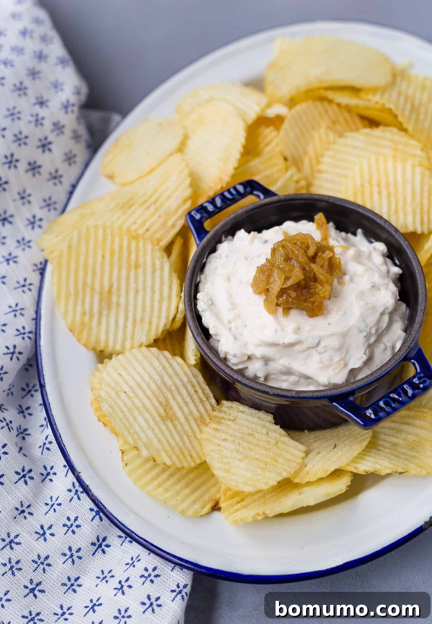 A plate of ridged potato chips arranged around a central bowl of creamy French onion dip, garnished with caramelized onions, next to a stylish patterned cloth.