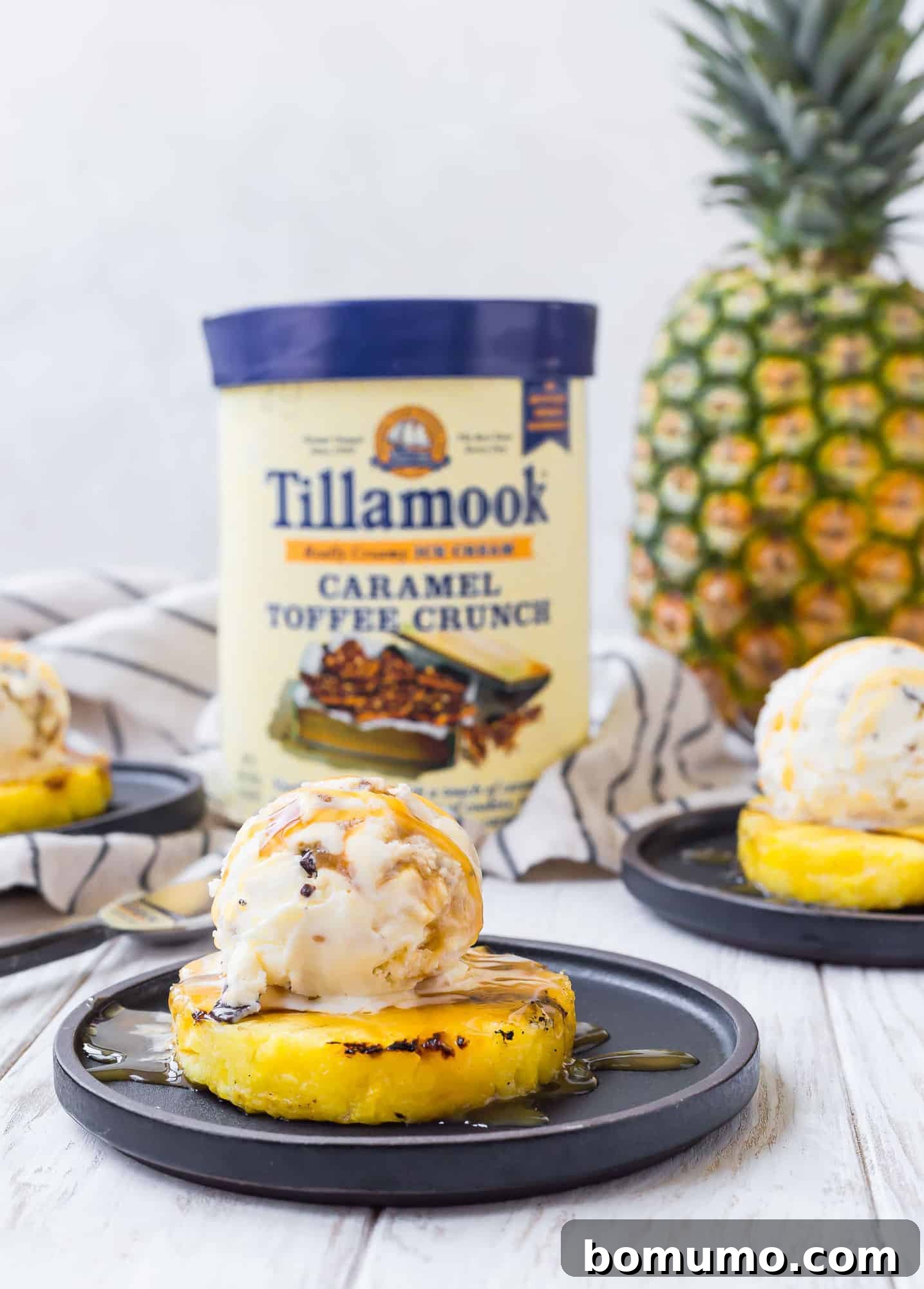 A beautifully presented slice of grilled pineapple with ice cream in the sharp foreground, with a familiar carton of Tillamook ice cream subtly blurred in the background, creating an enticing visual narrative of sweet indulgence.