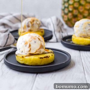 Grilled pineapple on a sleek black plate, elegantly topped with a generous scoop of ice cream and exquisitely drizzled with rich caramel sauce, capturing its delicious appeal.