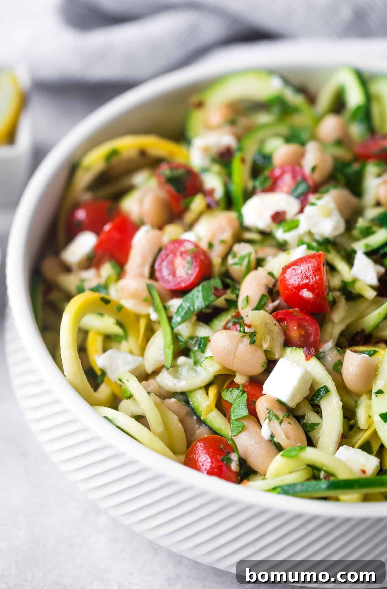 Close-up of zucchini noodles in a white bowl with fresh herbs, halved grape tomatoes, and black beans.