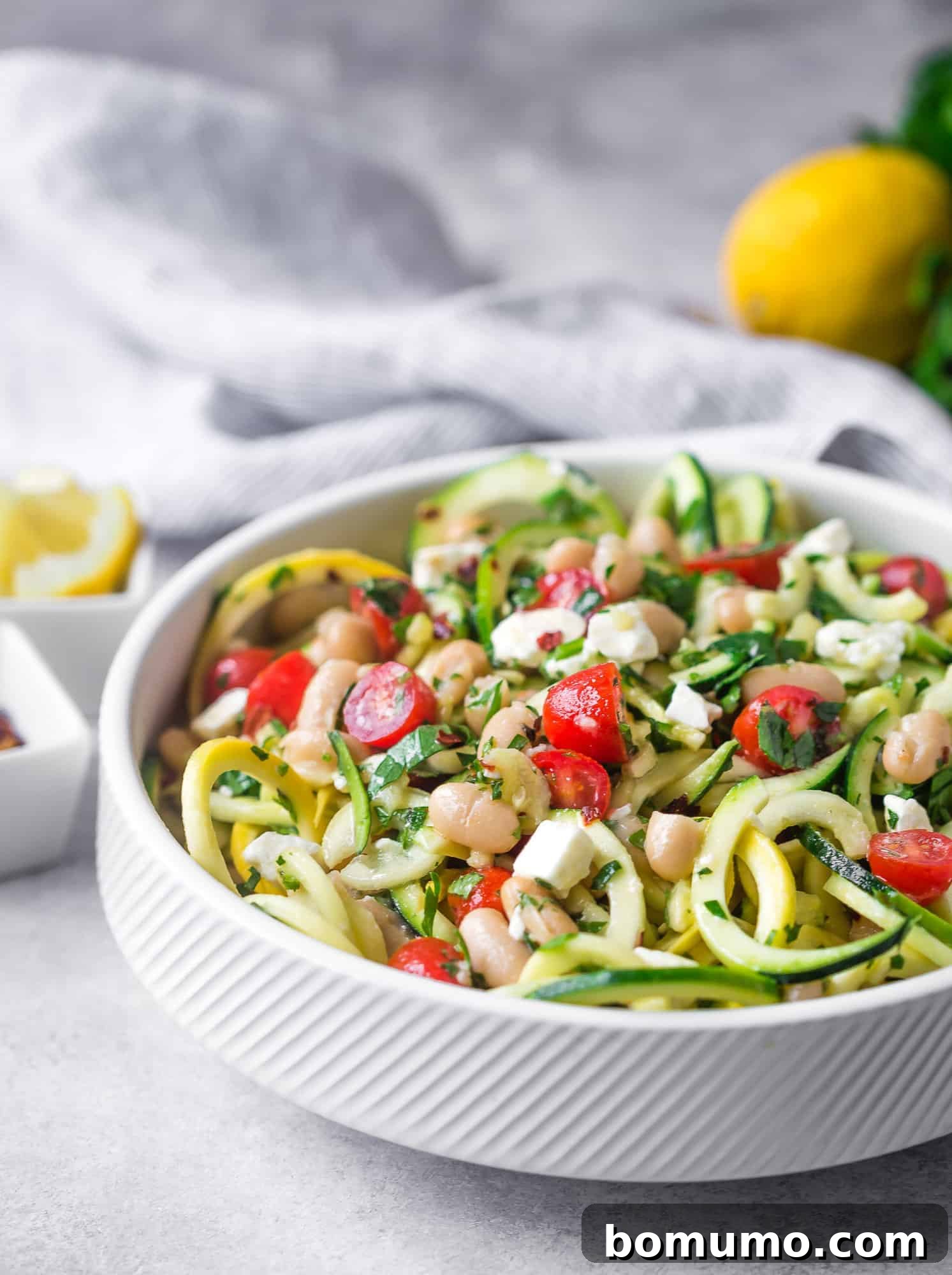 Zucchini noodle salad served in a white bowl, highlighted with tomatoes, feta cheese, and beans.