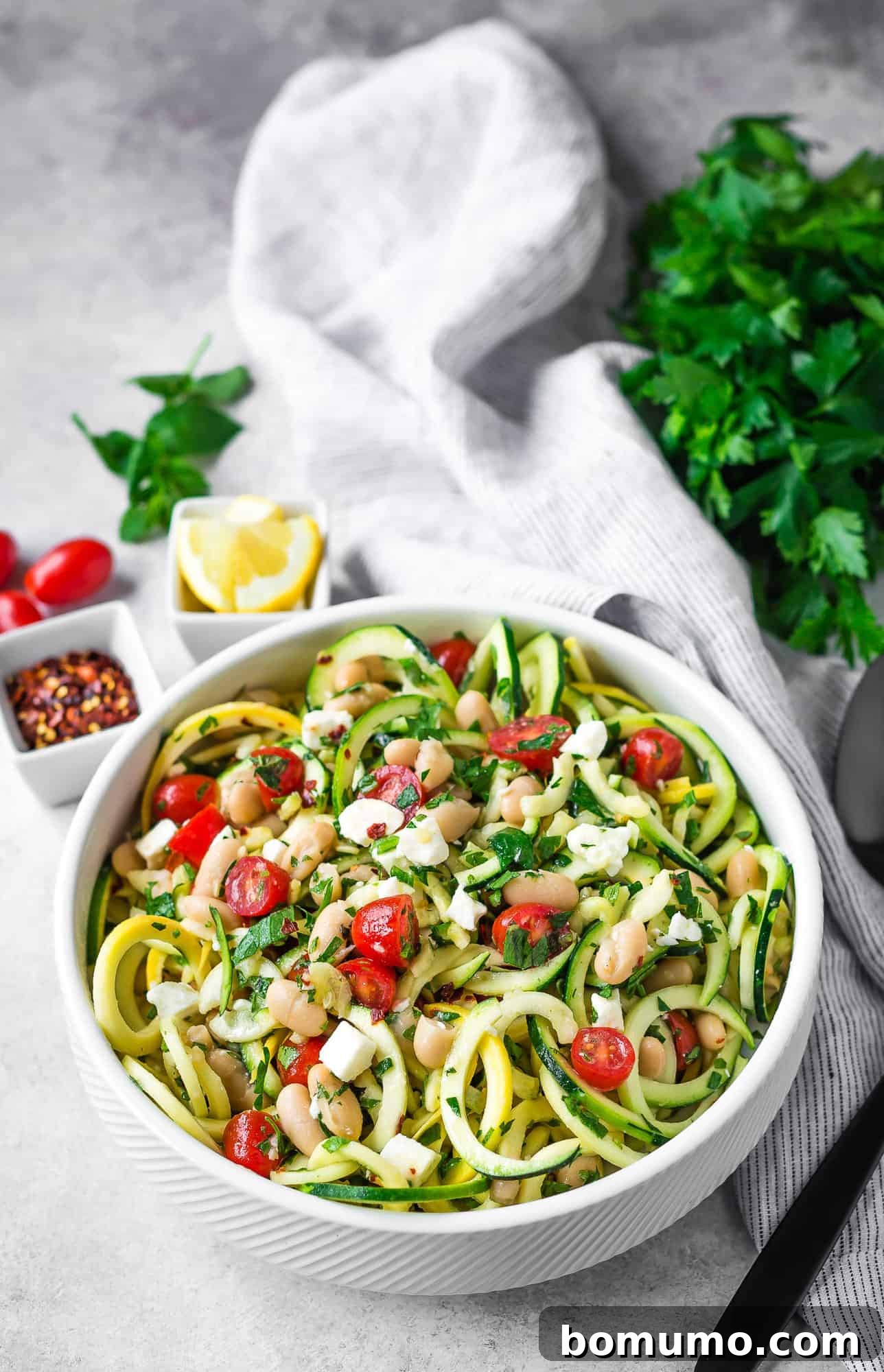 A shallow white bowl filled with zucchini noodle salad, surrounded by scattered fresh ingredients like tomatoes and herbs.