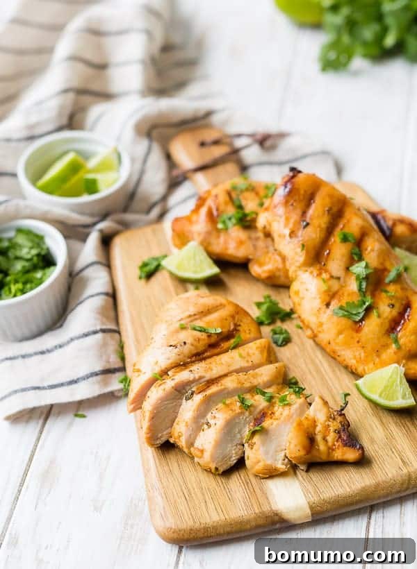 A sliced, grilled chicken breast is on a wooden cutting board with two other chicken breasts. Lime wedges are also pictured.