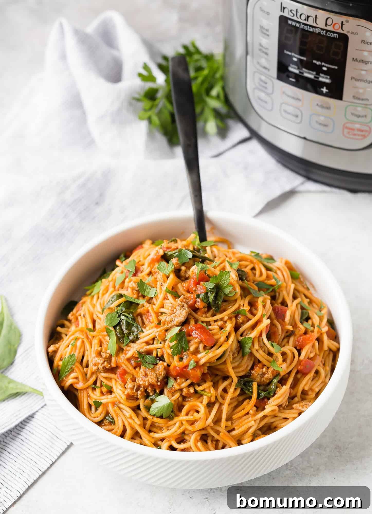A large white serving bowl filled with perfectly cooked Instant Pot spaghetti in a rich tomato and meat sauce, with a pressure cooker and fresh parsley in the blurred background, emphasizing the one-pot convenience.