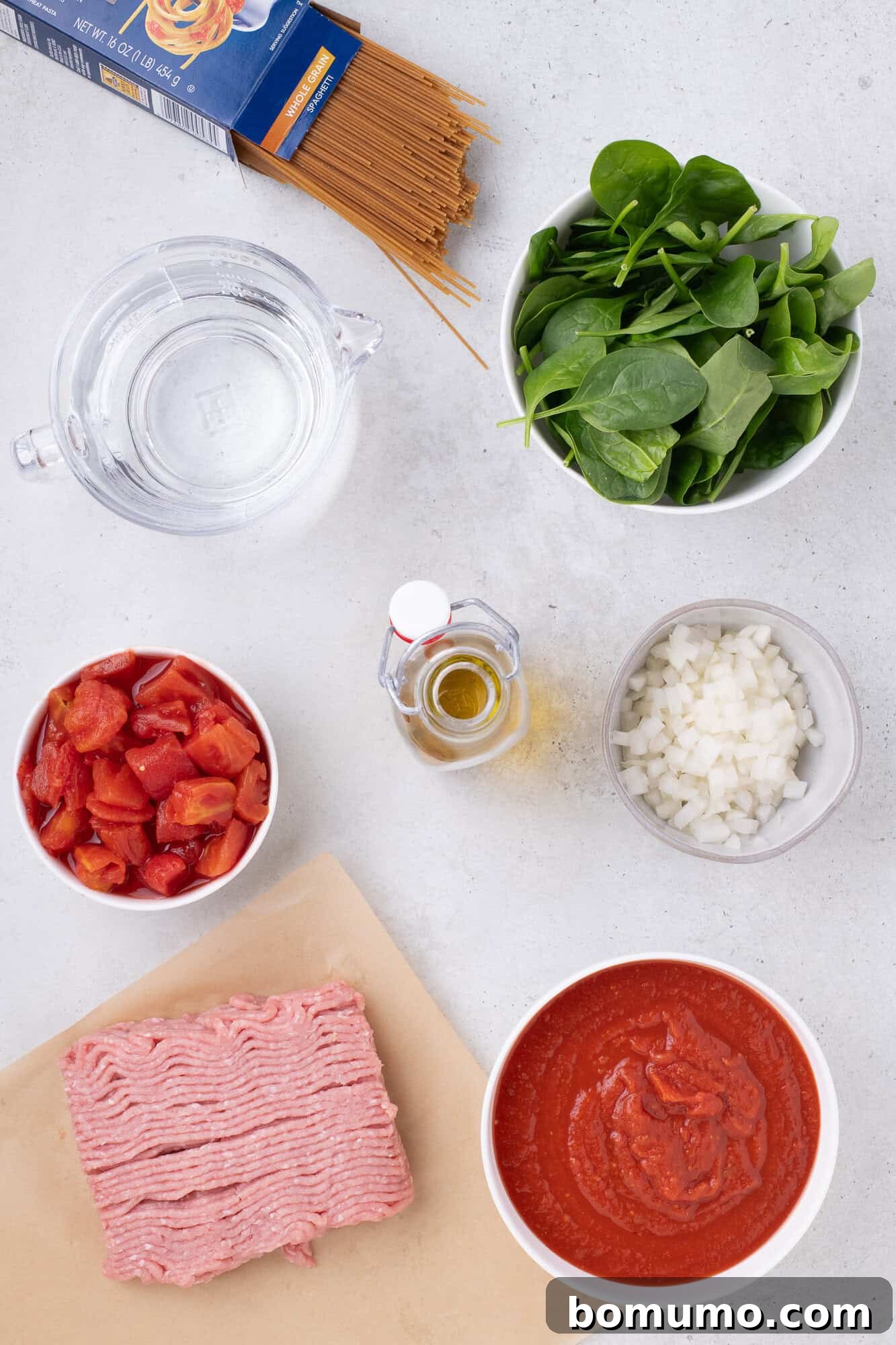 Overhead view of fresh ingredients meticulously arranged for the Instant Pot Spaghetti recipe, including ground turkey, olive oil, diced onions, uncooked spaghetti, jarred sauce, canned diced tomatoes, and a pile of fresh spinach leaves.