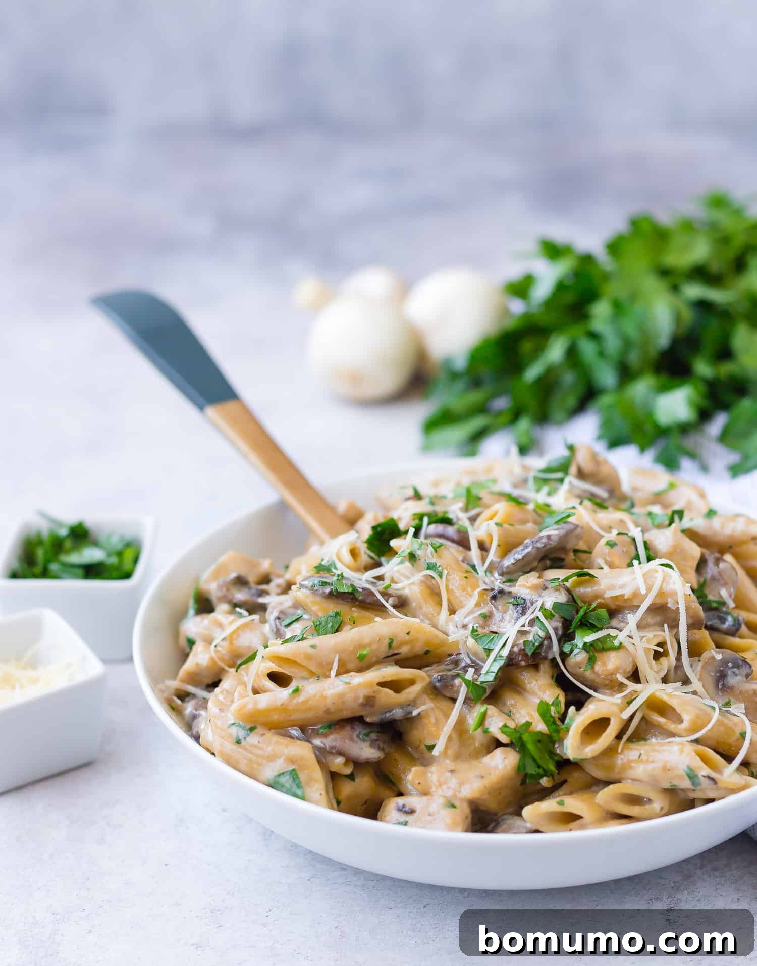 Pasta in serving bowl with mushrooms and parsley in background.