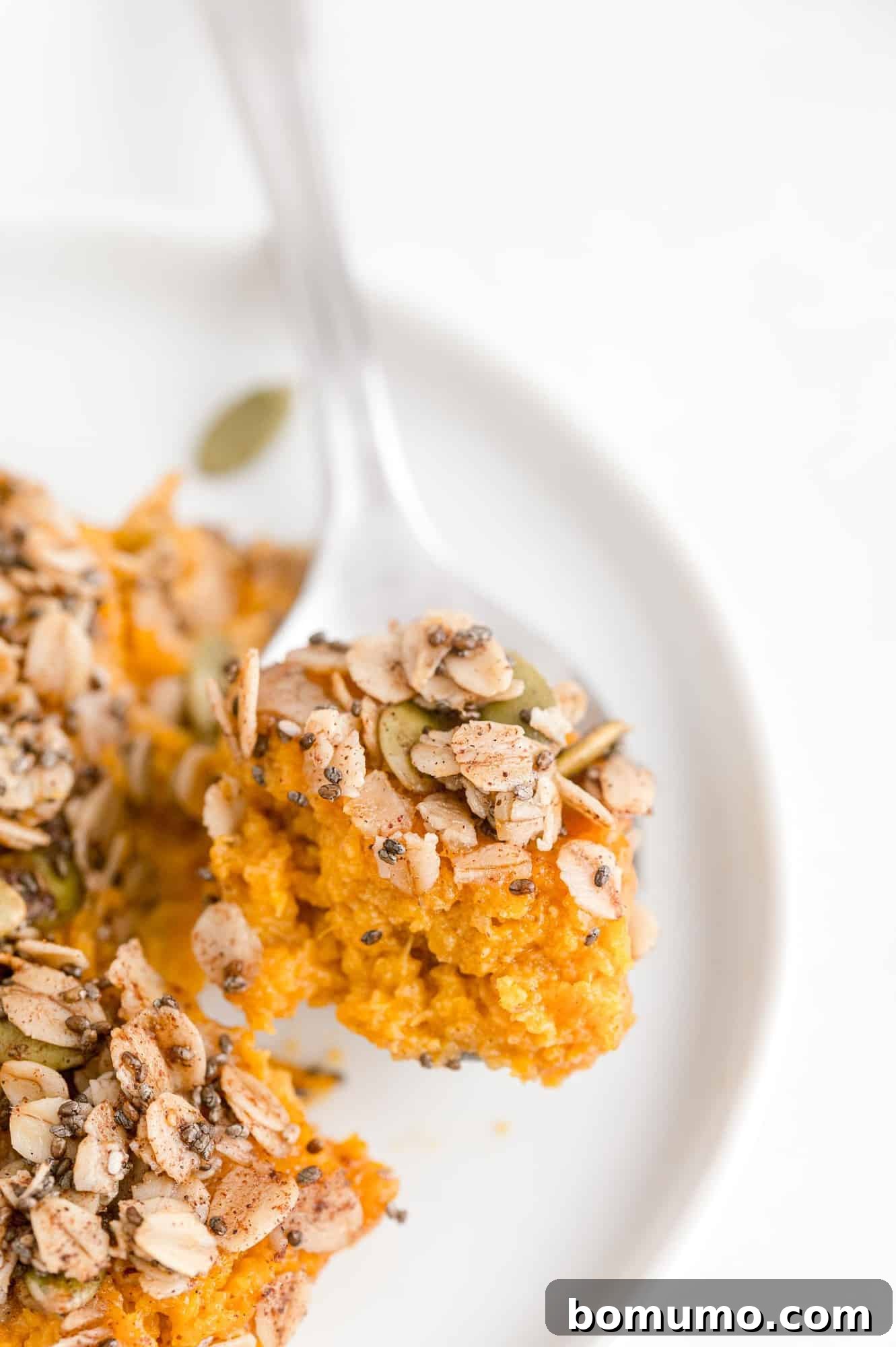 A close-up view of a perfect bite of healthy sweet potato casserole, complete with its crunchy topping, delicately balanced on a spoon on a white plate.
