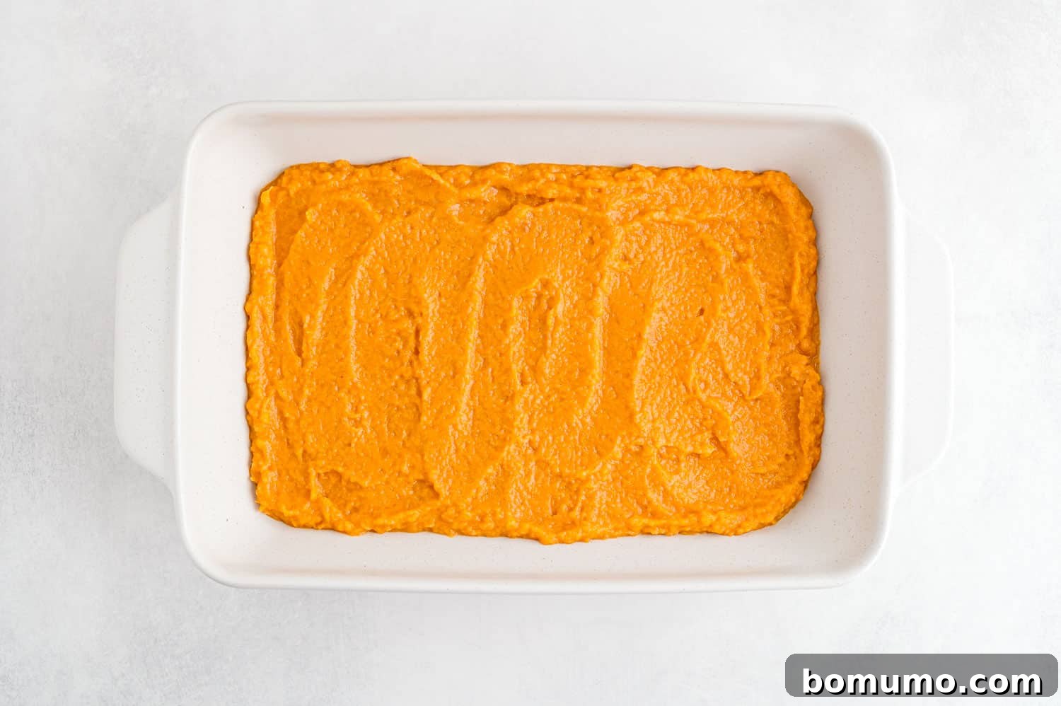 Creamy mashed sweet potatoes meticulously spread into an even, smooth layer within a pristine white casserole dish, awaiting its topping.
