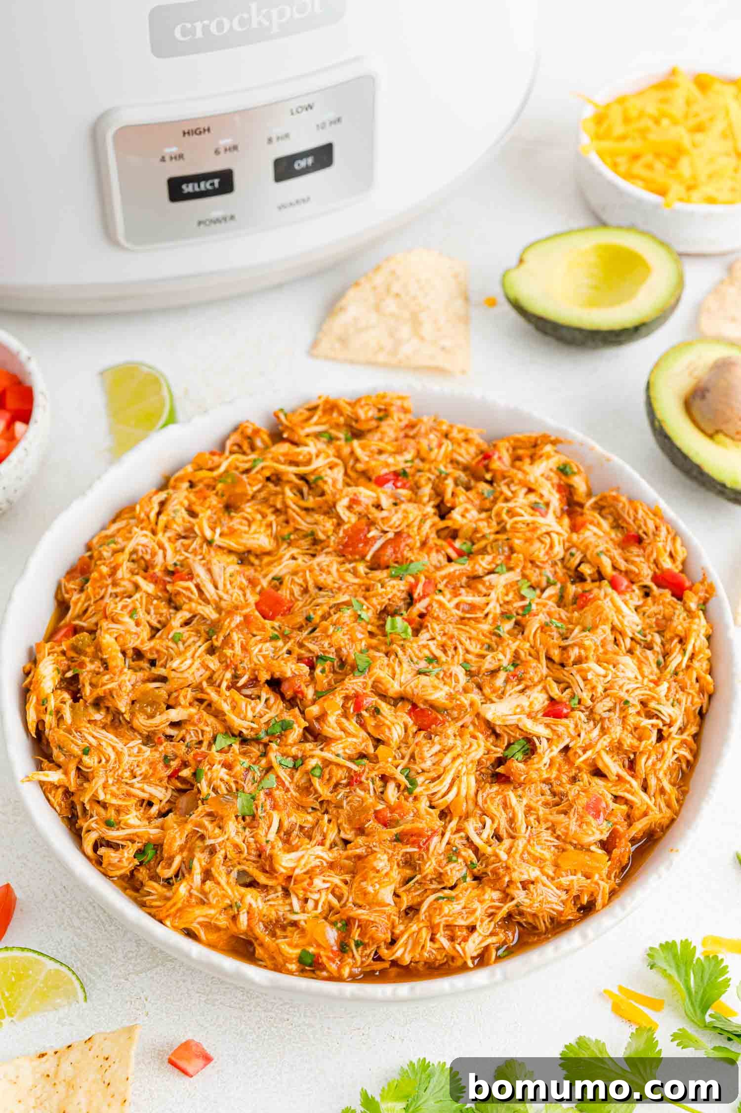 A large bowl of crockpot salsa chicken on a countertop next to the crockpot, surrounded by garnishes.
