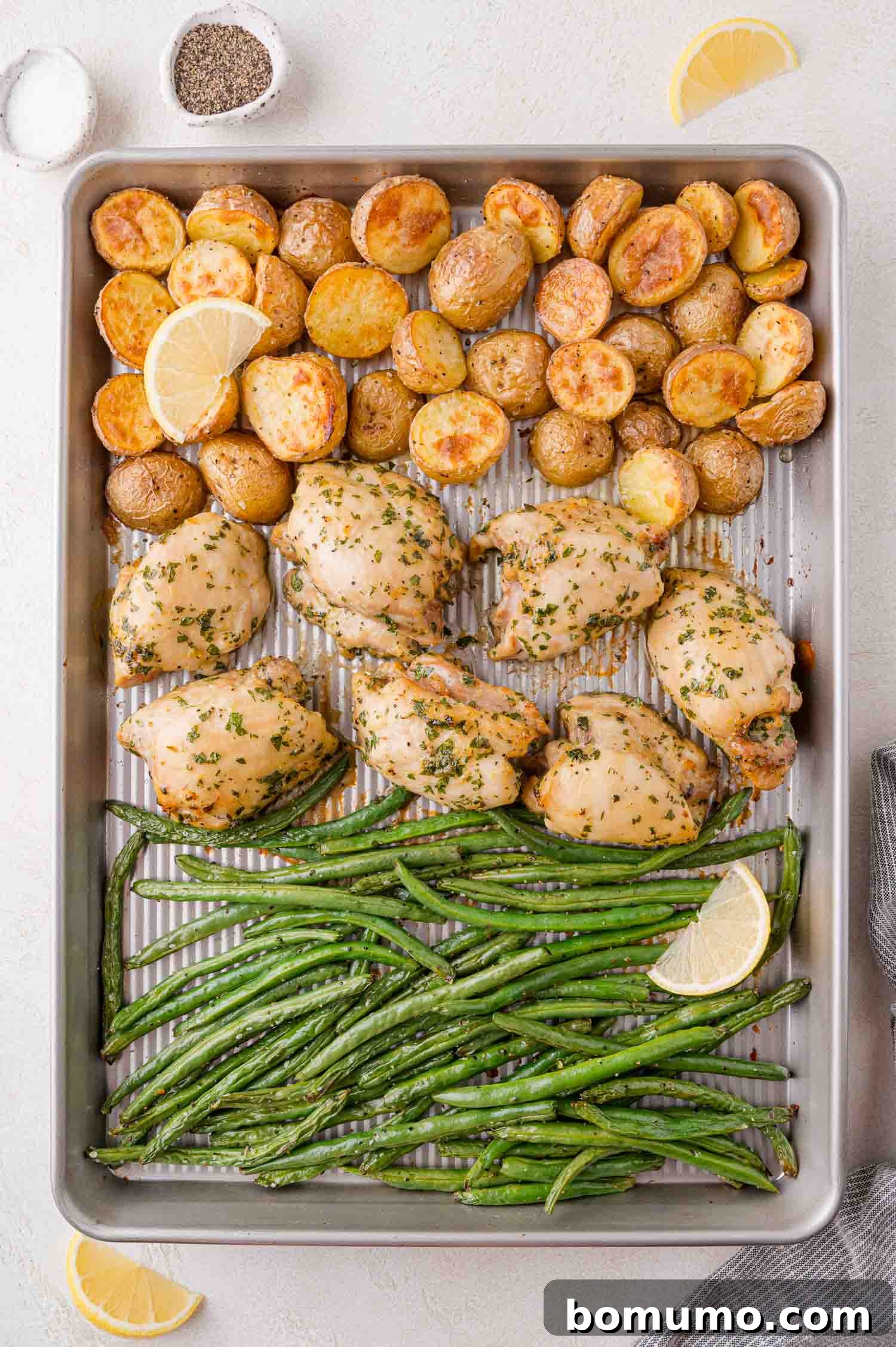 Sheet pan with roast chicken thighs, halved baby potatoes, and green beans garnished with lemon slices.