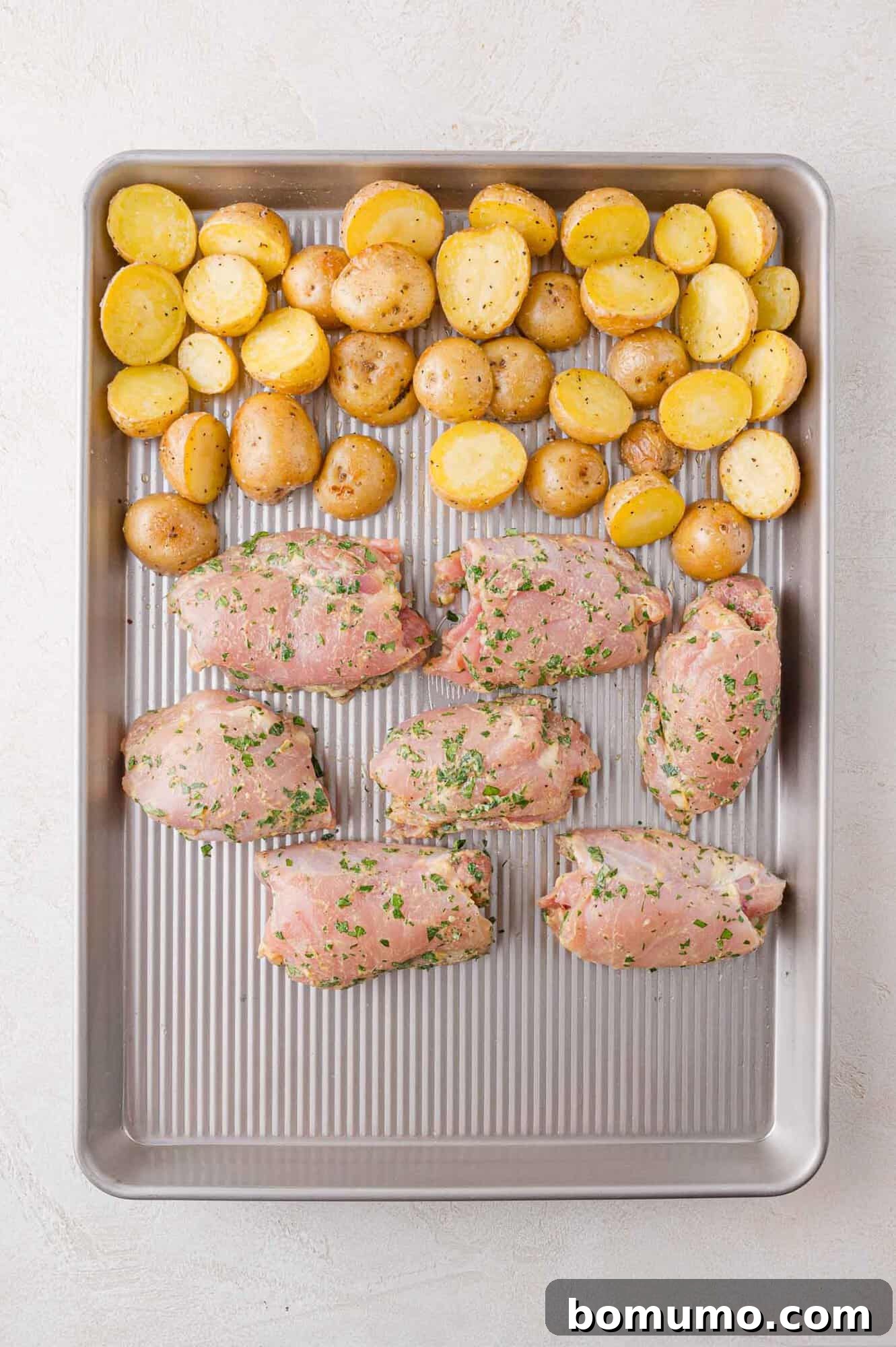 A baking tray with seasoned chicken and halved potatoes ready for roasting.