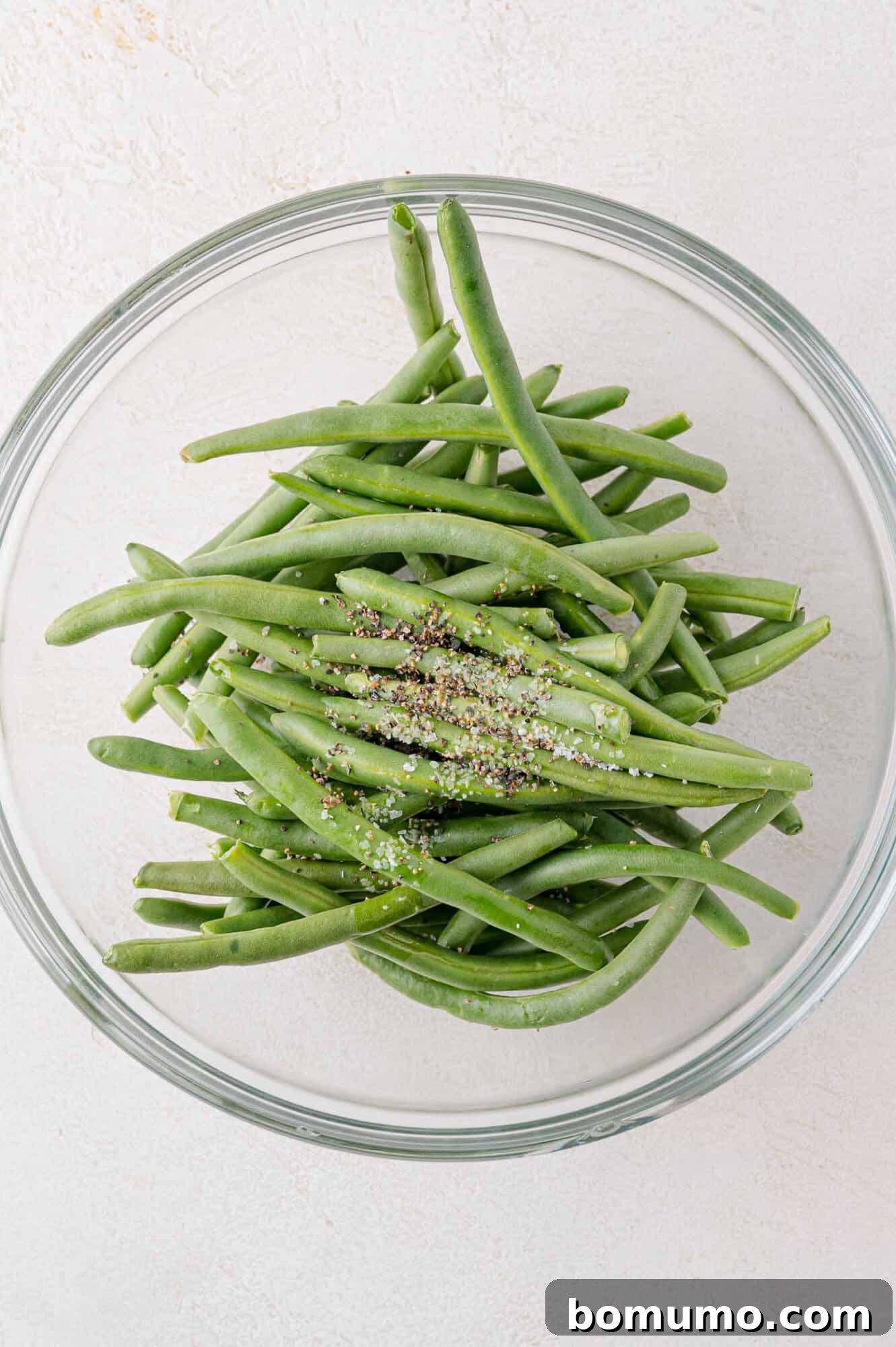A clear glass bowl filled with fresh green beans, topped with a sprinkle of black pepper and sea salt, on a light-colored surface.
