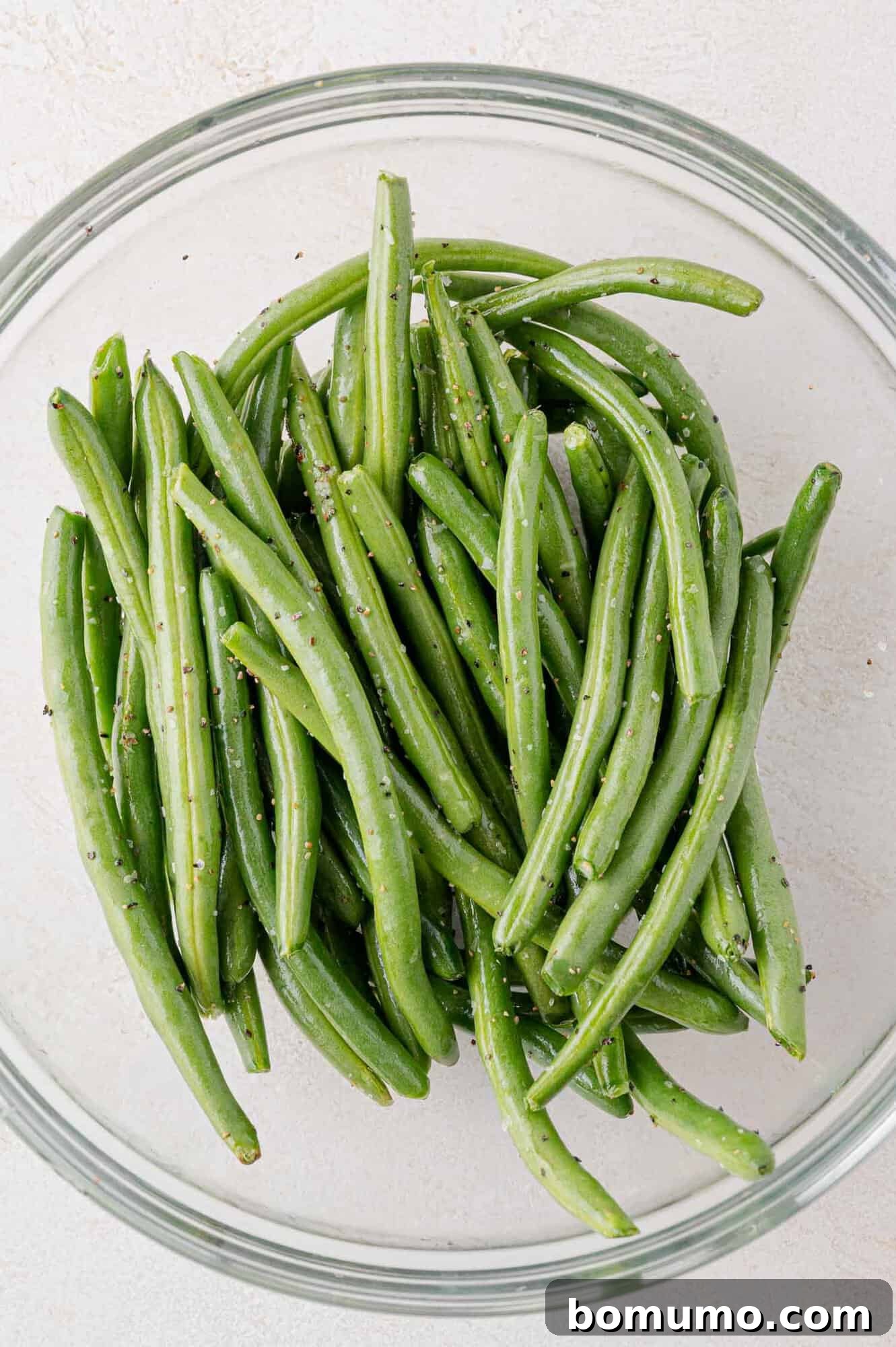 A clear glass bowl containing fresh green beans seasoned with salt and pepper on a light surface.