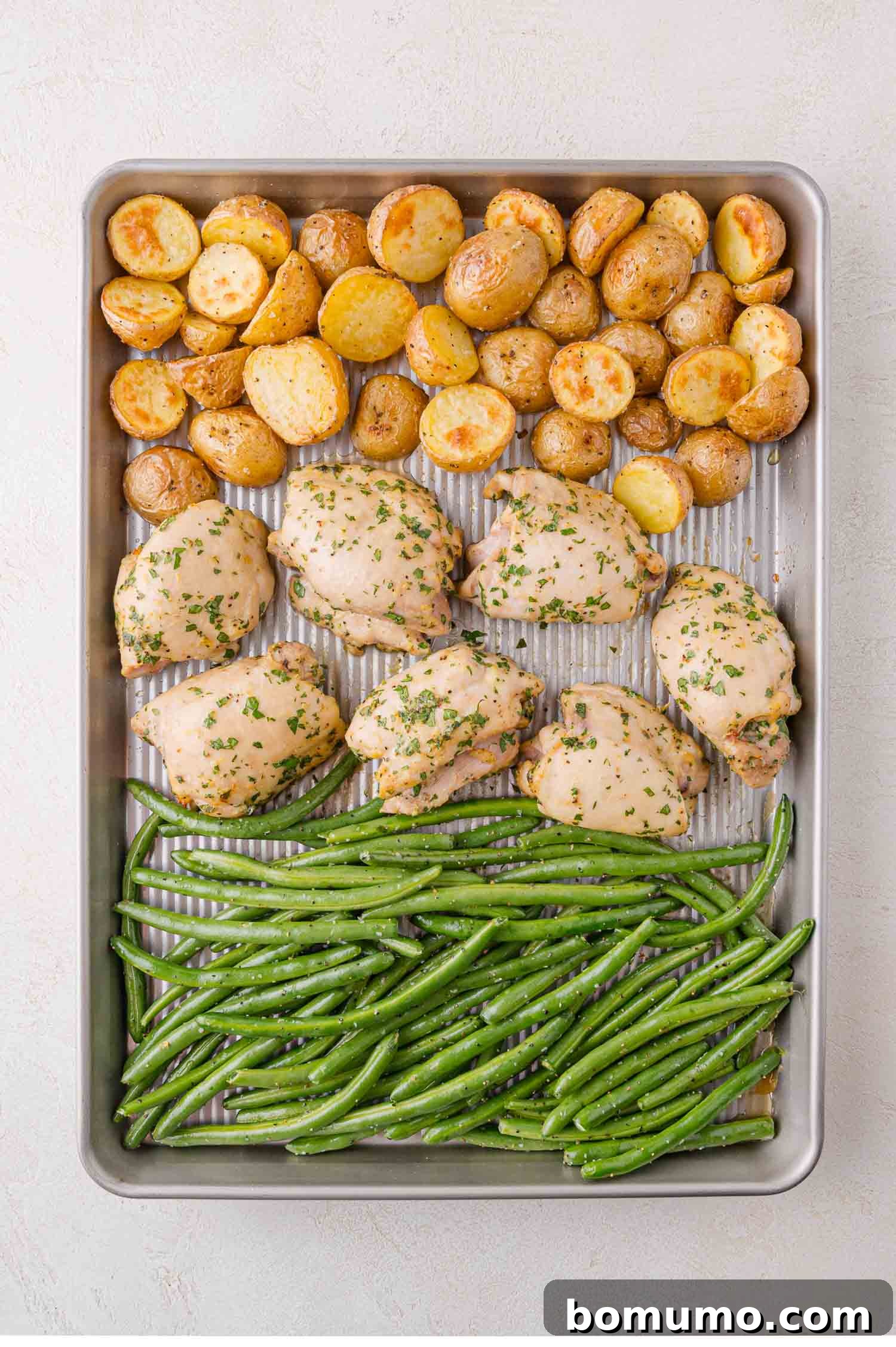 Sheet pan with roasted potatoes, seasoned chicken thighs, and green beans arranged in rows on a metal baking tray.
