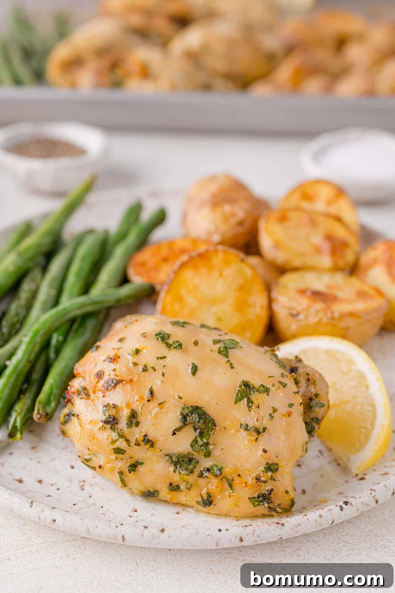 Plate of herb-marinated chicken thigh with roasted potatoes, green beans, and a lemon wedge. Salt and pepper shakers are in the background.