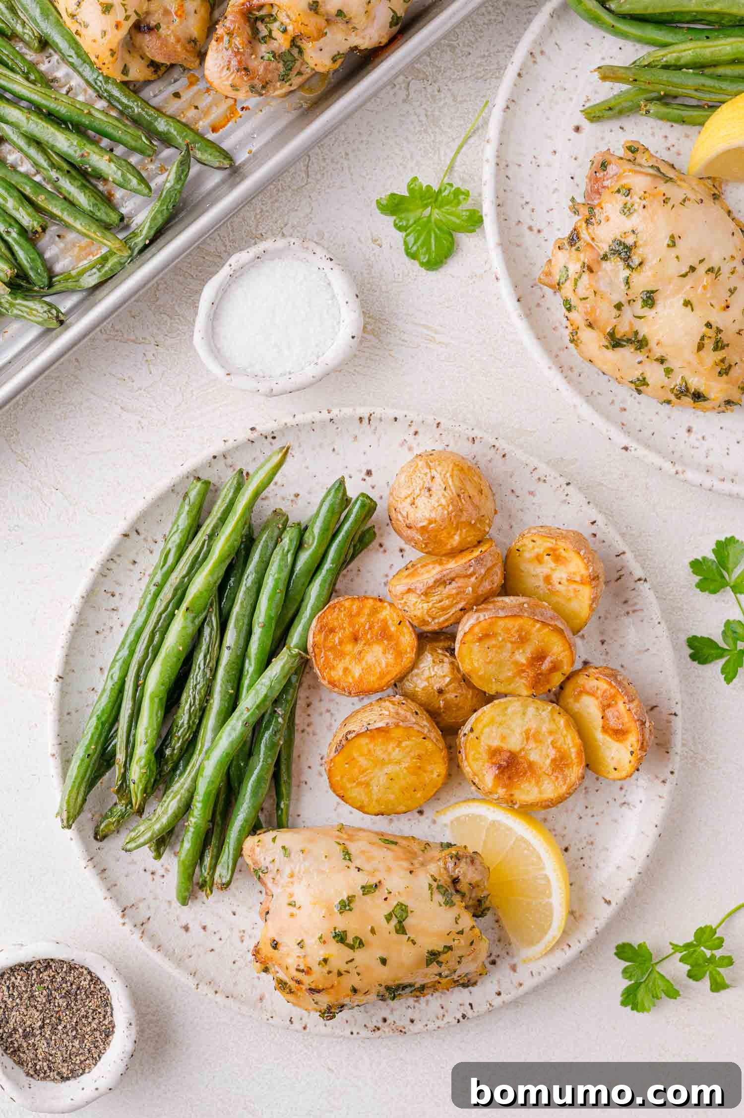 A plate with roasted chicken, green beans, and sliced potatoes. Next to the plate are dishes of pepper and salt. A lemon wedge and parsley garnish the meal.