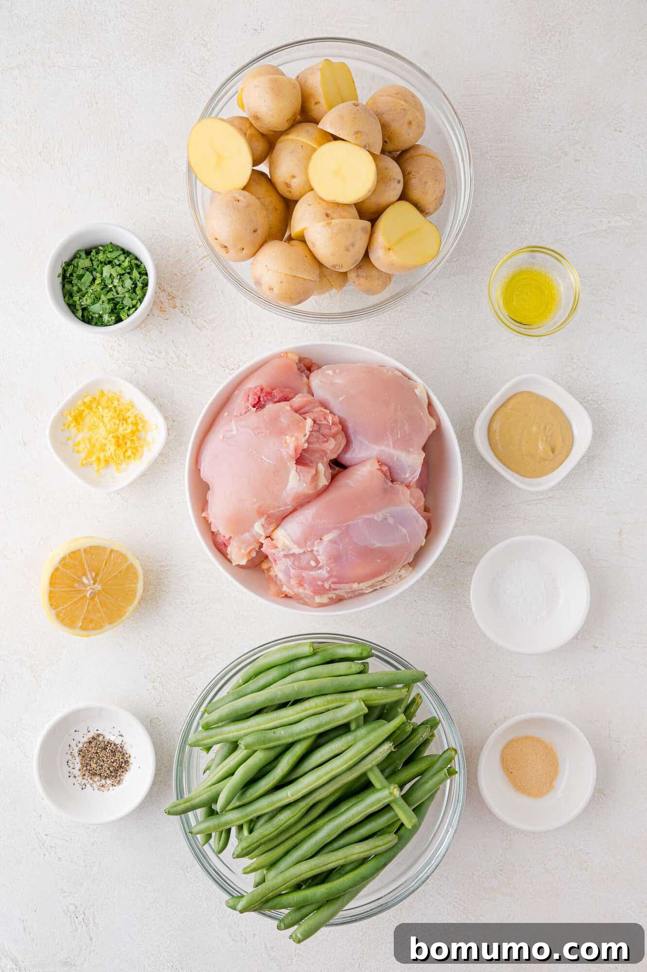 Bowls of raw chicken thighs, green beans, and potatoes with small bowls of lemon, mustard, oil, seasonings, and chopped herbs on a white surface.