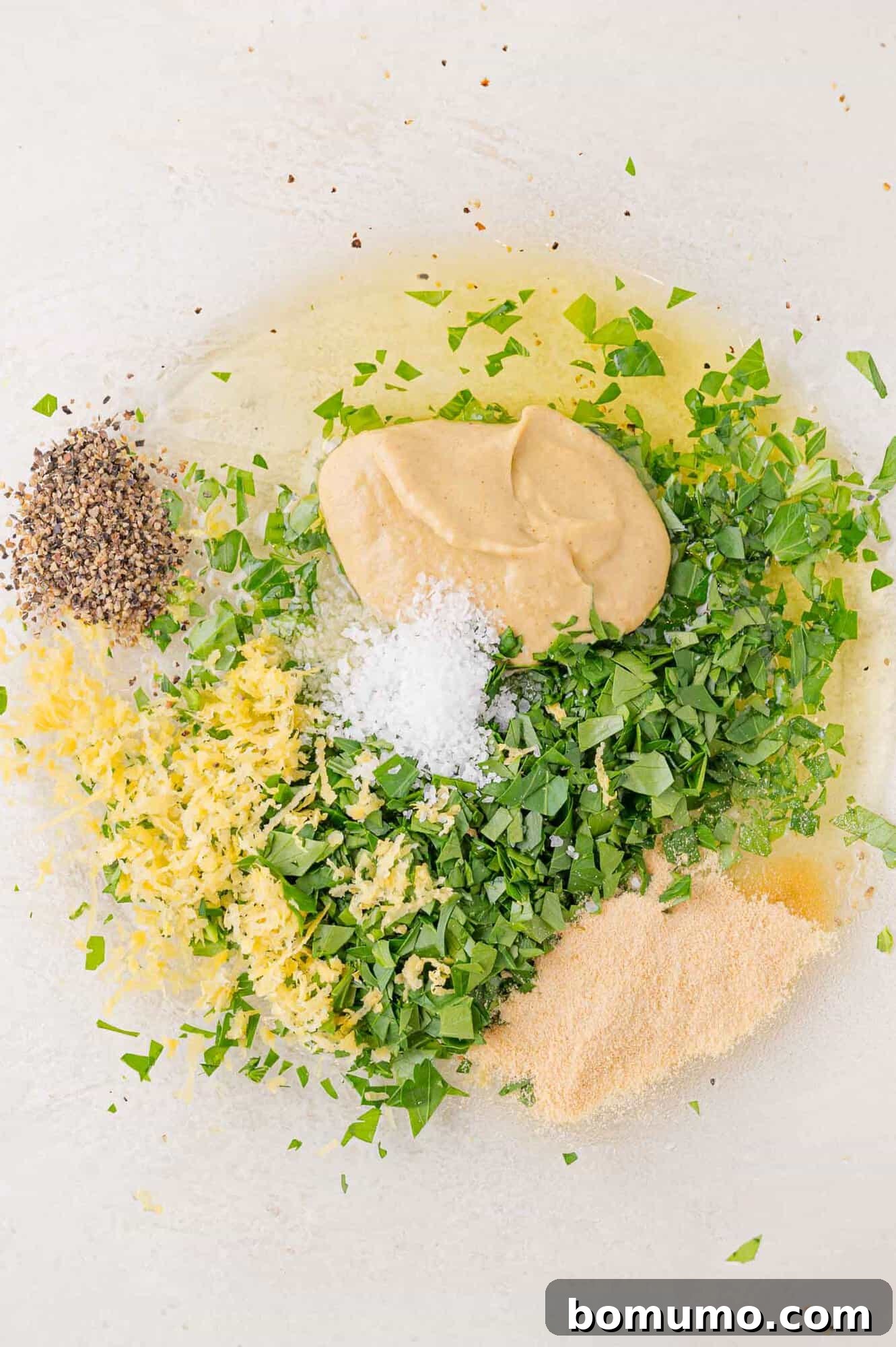 Ingredients for a dressing: mustard, parsley, lemon zest, cracked black pepper, salt, and oil on a white surface.