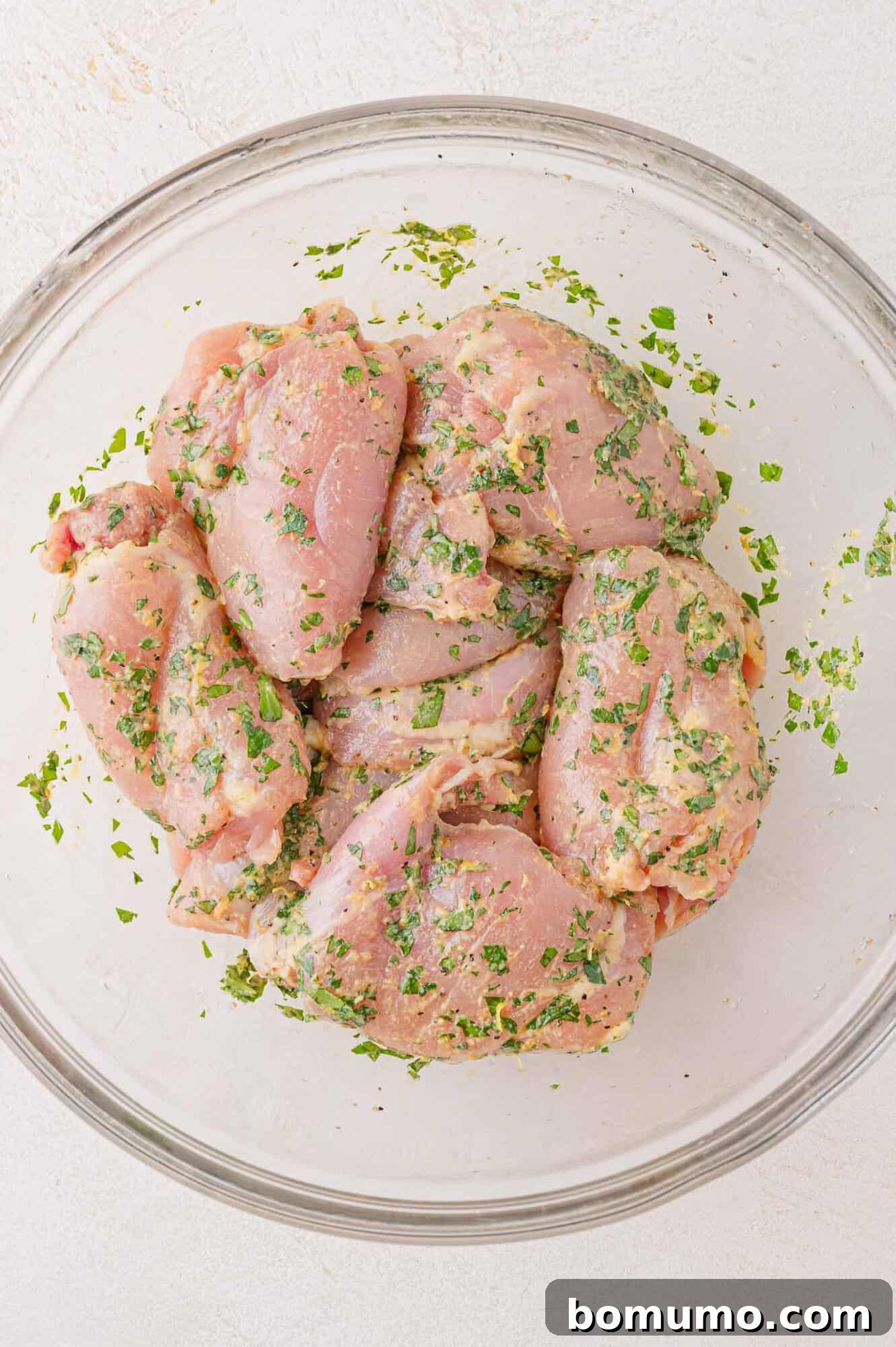 Raw chicken pieces marinated with herbs and spices in a clear glass bowl on a light background.
