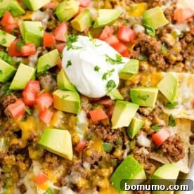 Close up view of a tray of beef nachos topped with cheese, diced tomatoes, avocados, onions, and dollops of sour cream.