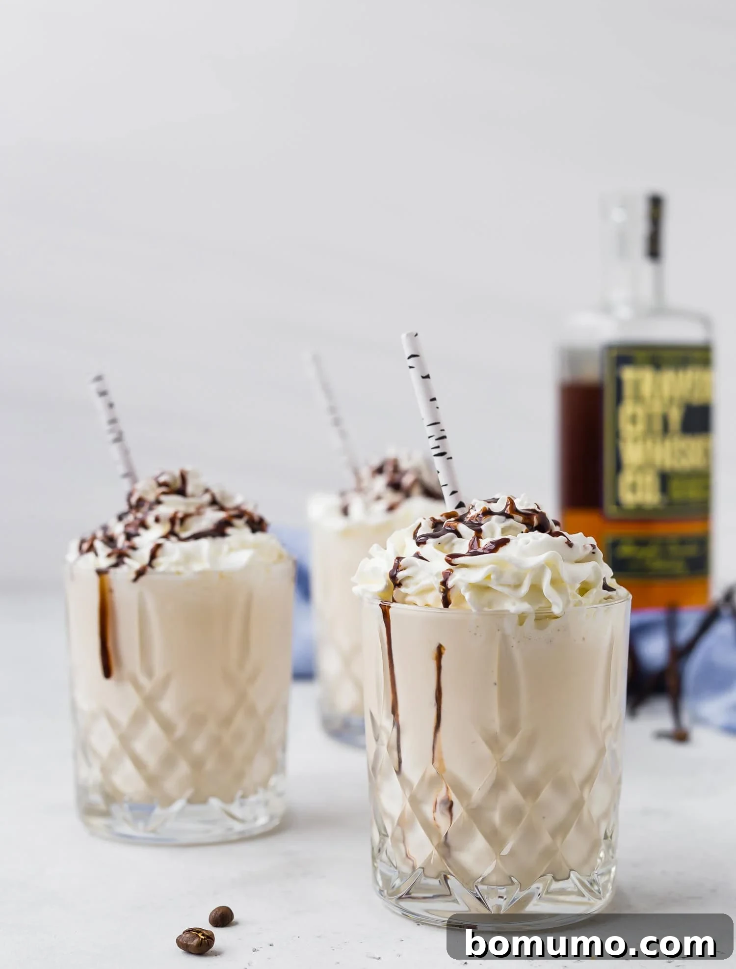 Indulge in the best Maple Whiskey Milkshake with creamy whipped topping and decadent chocolate drizzle. Three whiskey milkshakes topped with whipped cream and chocolate drizzle, served in patterned glasses with striped straws. A bottle of alcohol is in the background.