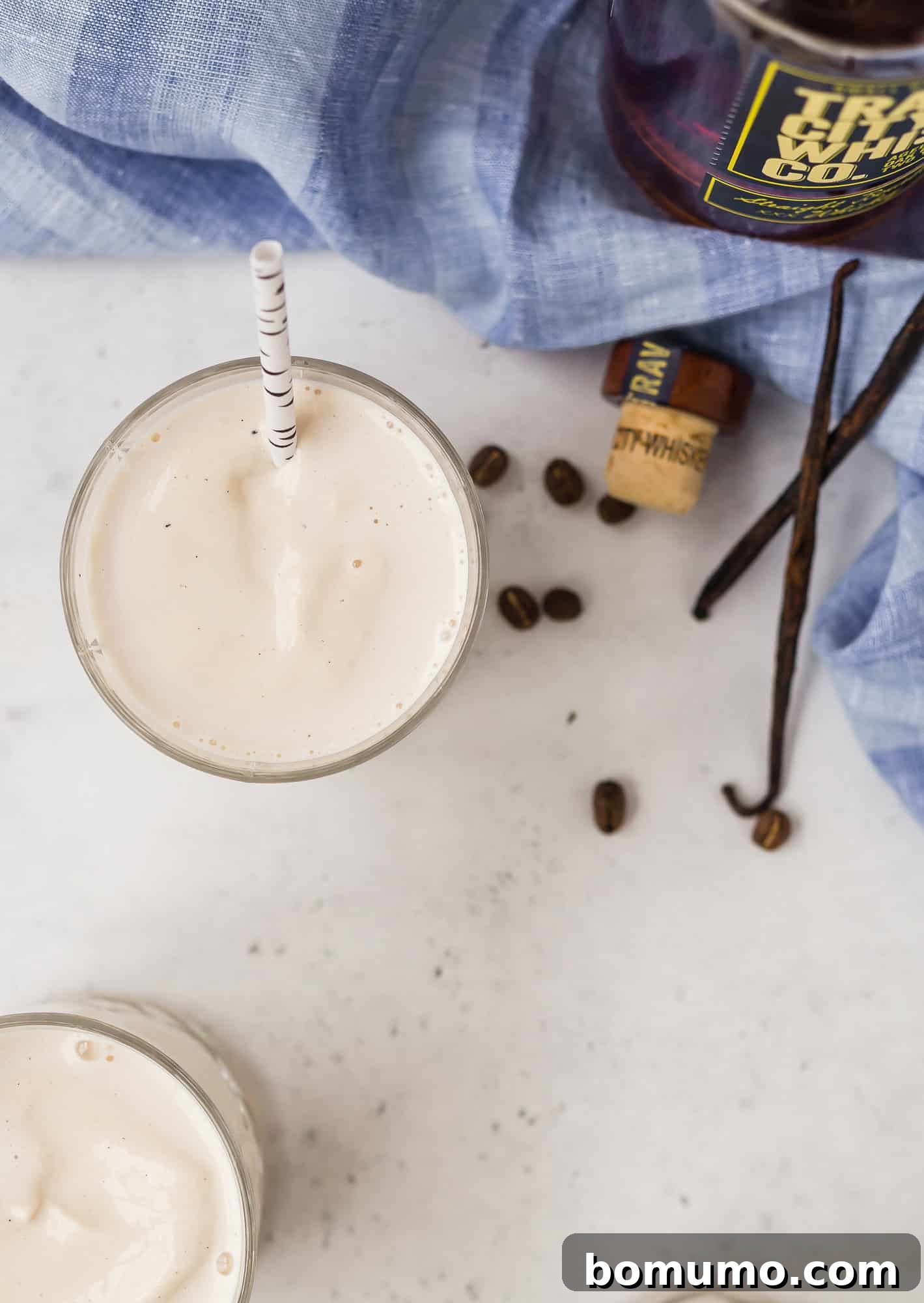 Creamy Maple Whiskey Milkshake adorned with coffee beans and vanilla for an aromatic touch. A creamy milkshake with a straw sits on a white surface near coffee beans, a cork, vanilla beans, and a bottle, with a blue cloth partially visible.