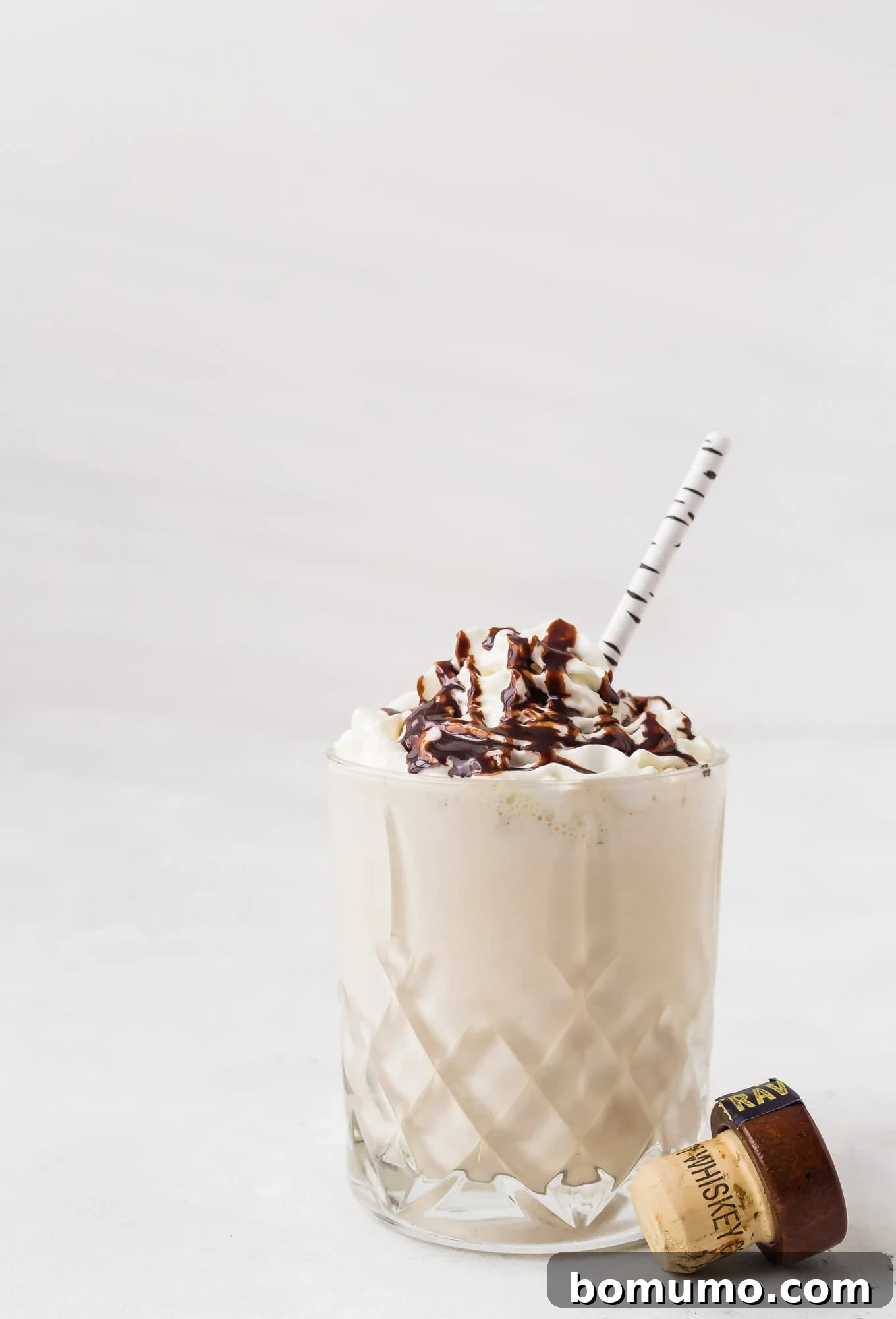 Perfectly garnished Maple Whiskey Milkshake with whipped cream and chocolate syrup, ready to enjoy. A glass of creamy milkshake topped with whipped cream and chocolate drizzle, with a striped straw and a bottle cap beside the glass.