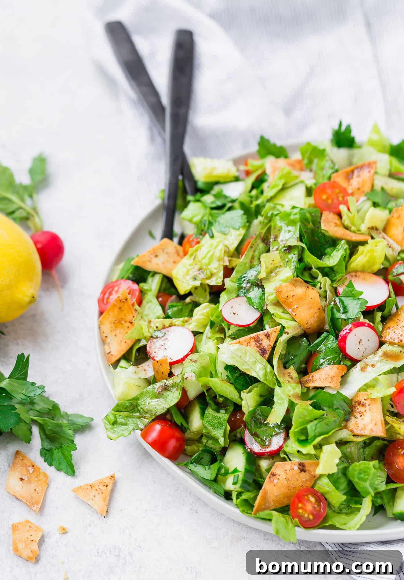 A large serving platter filled with vibrant Fattoush salad, featuring crisp romaine lettuce, halved cherry tomatoes, thinly sliced radishes, fresh parsley, and golden-brown crispy pita pieces. Black serving utensils are resting beside the platter.