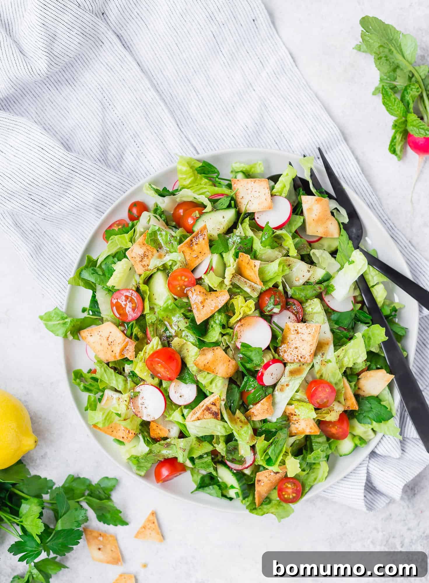 A vibrant Fattoush salad arranged on a large serving platter, artfully garnished with fresh herbs and golden-brown toasted pita bread. Black serving utensils are placed alongside the dish, ready for serving.