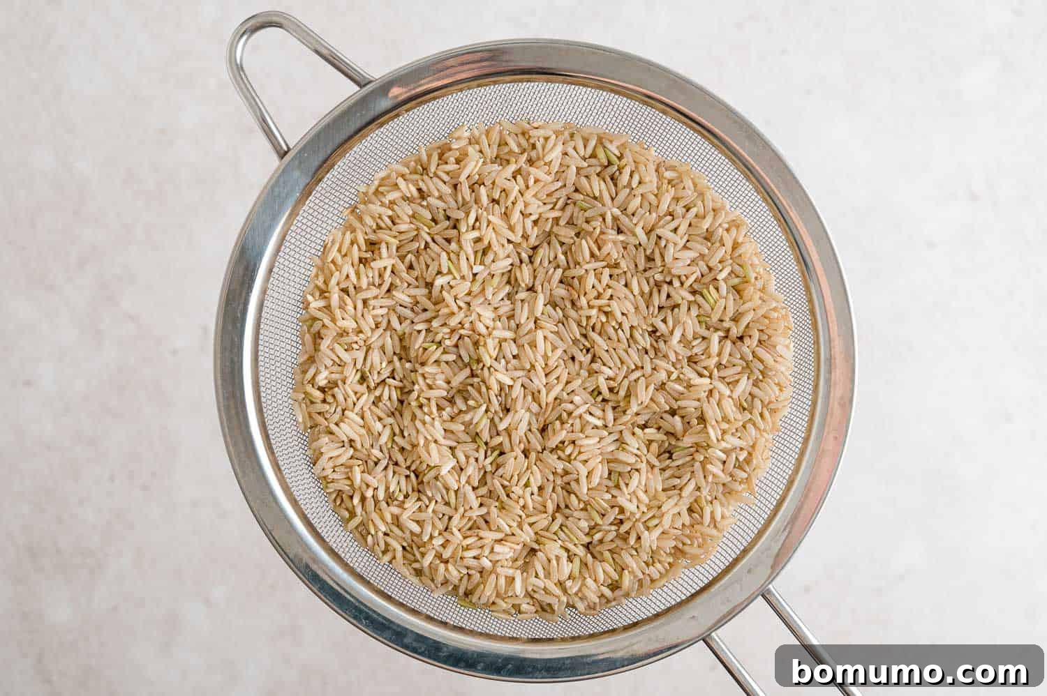 Raw brown rice being rinsed thoroughly under cold running water in a fine mesh strainer.