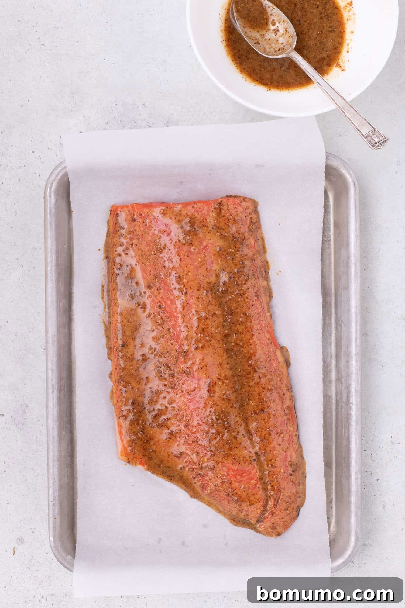 Maple mustard glaze generously added to an uncooked salmon fillet on a baking sheet.