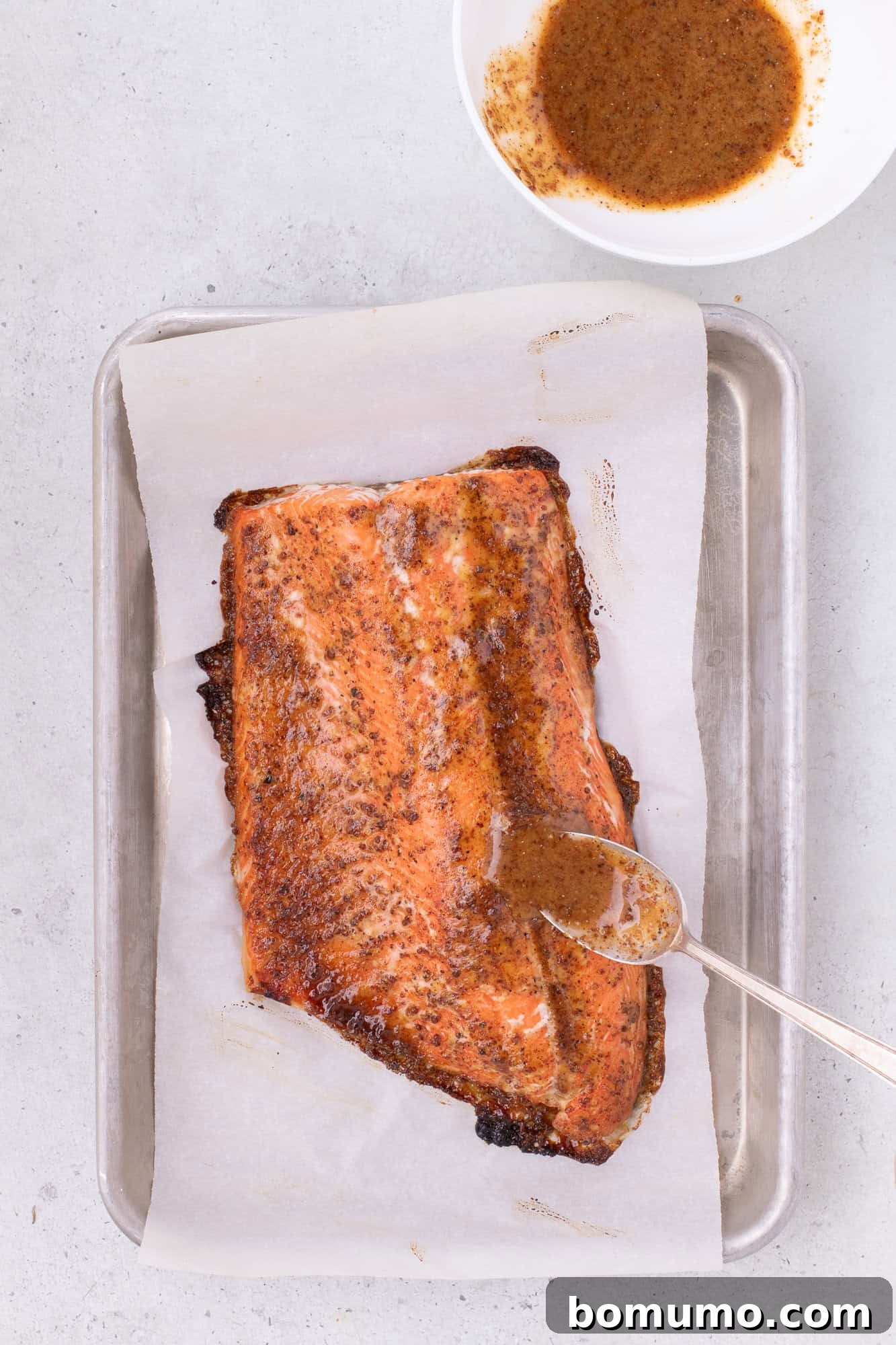 Additional glaze being added to a partially baked salmon fillet on a baking sheet, for a rich, caramelized finish.