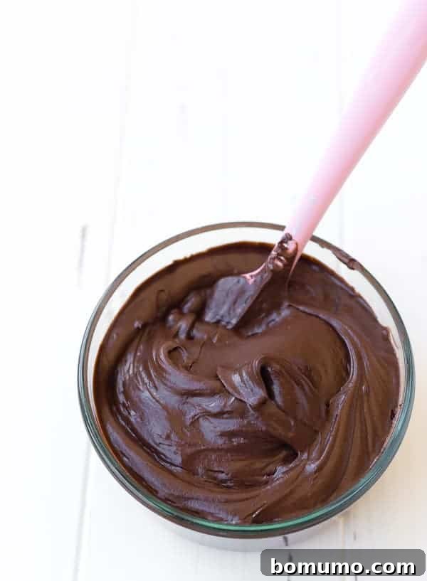Overhead view of a bowl filled with luscious chocolate cream cheese frosting, with a pink rubber scraper inserted.
