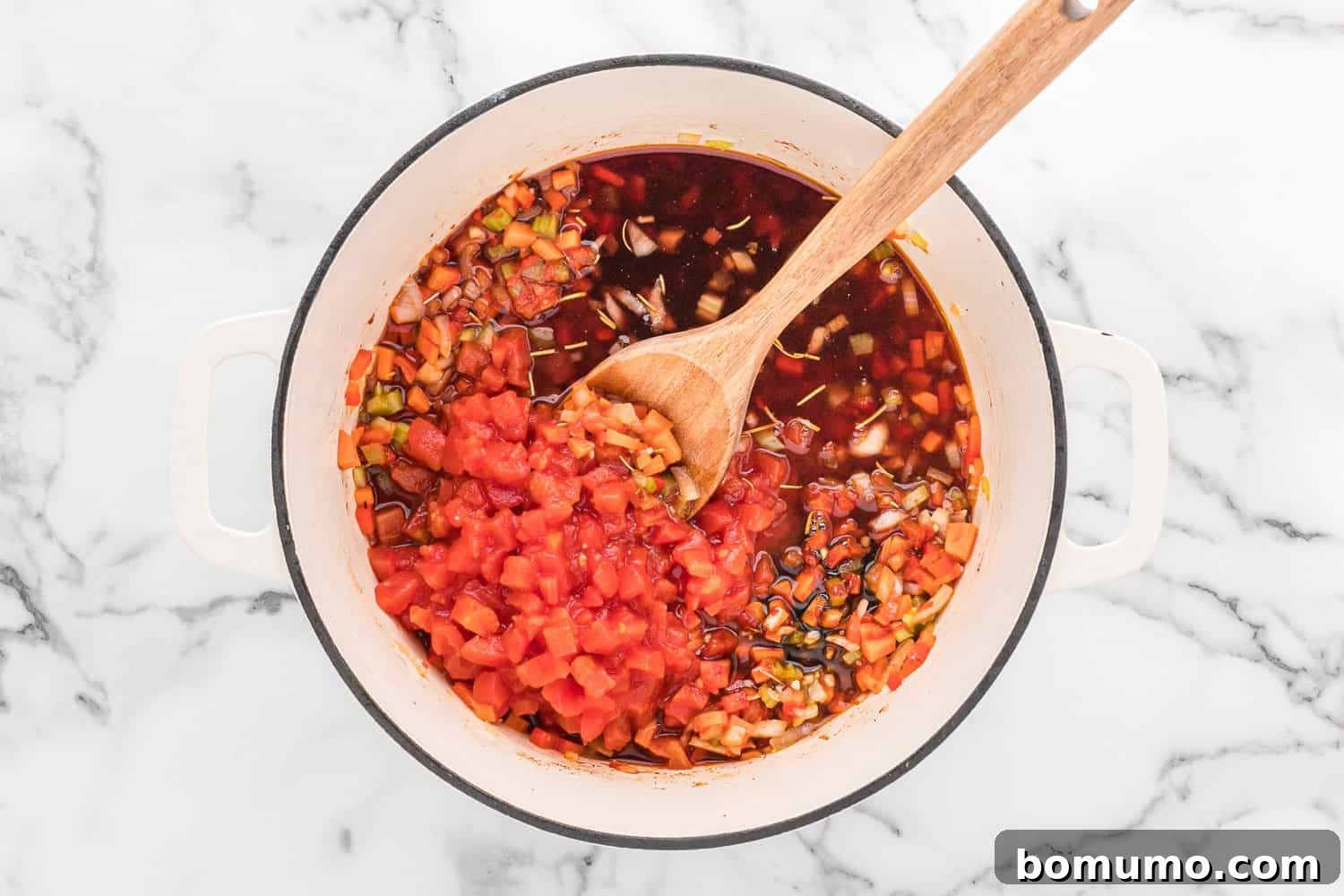 Canned diced tomatoes being added to the sautéed vegetables in a Dutch oven, deepening the sauce's flavor and color.