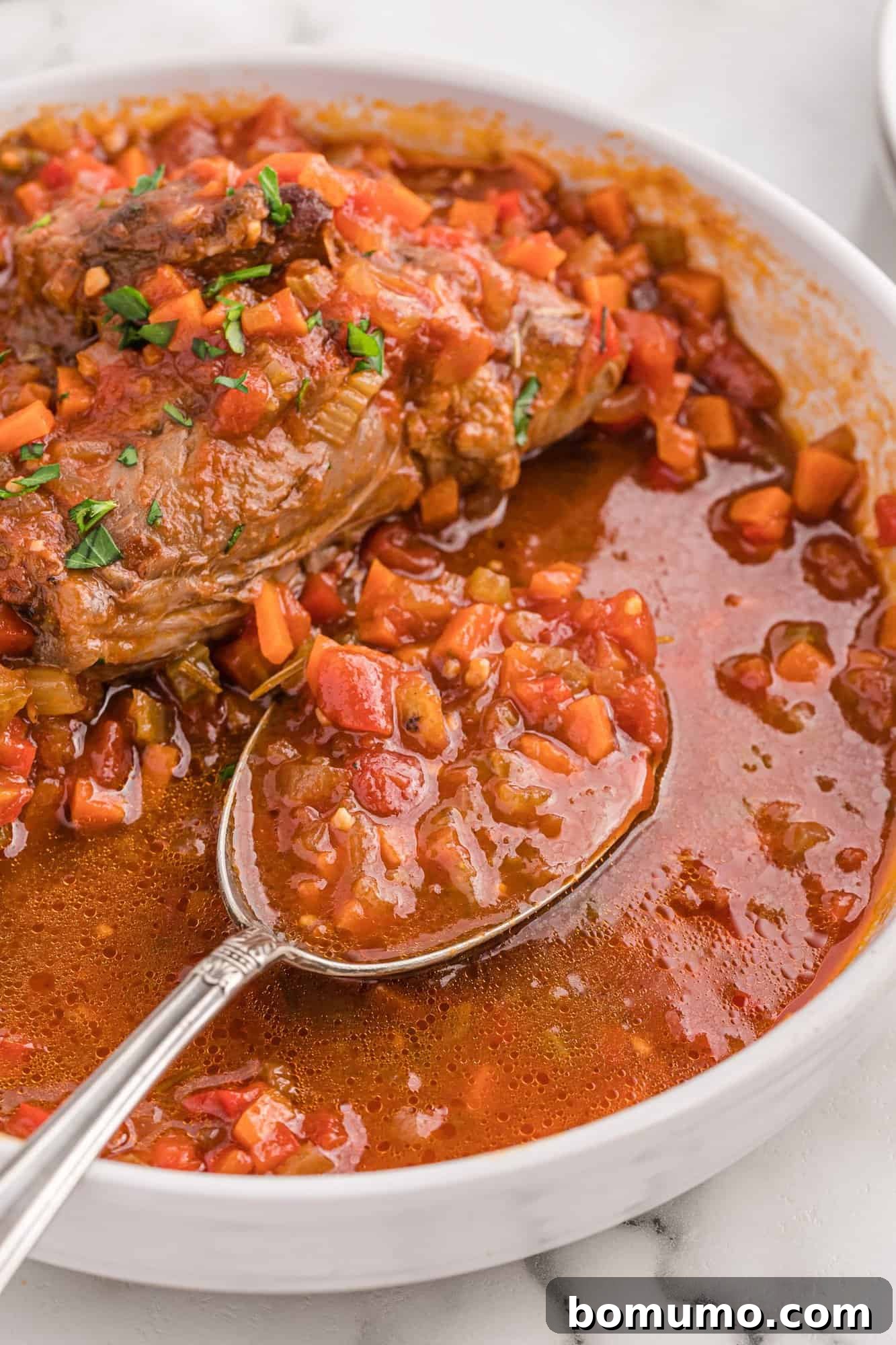 A large spoon lifting a generous portion of rich braising liquid, showcasing its thick consistency and flavorful depth, ready to accompany the tender lamb.