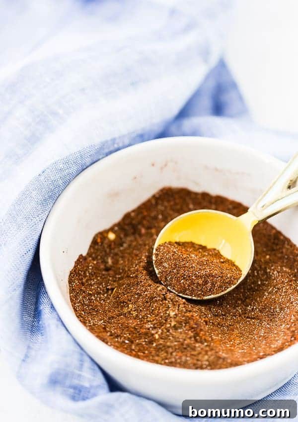 Close-up shot of various aromatic spices mixed together in a white bowl, ready to be used as chili seasoning, against a soft blue background.