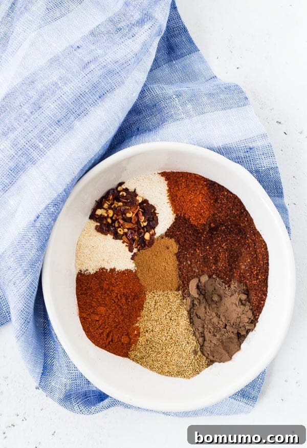 An overhead view of a selection of individual spices, neatly arranged in a white bowl, highlighting the various colors and textures before being mixed into a chili seasoning.