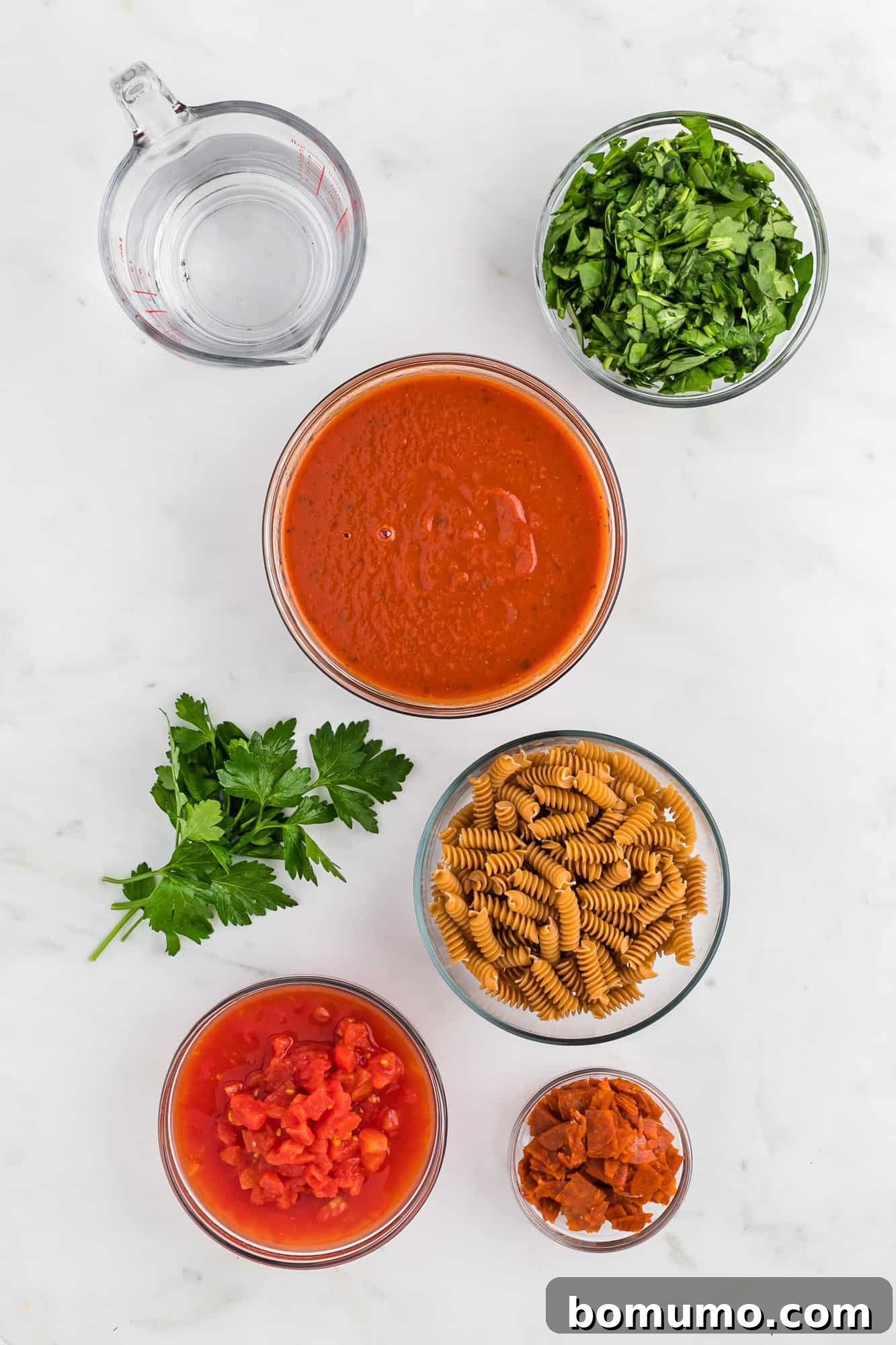 Overhead view of fresh ingredients for One-Pan Pizza Pasta laid out in separate bowls: pasta, pizza sauce, diced tomatoes, pepperoni, spinach, and mozzarella.