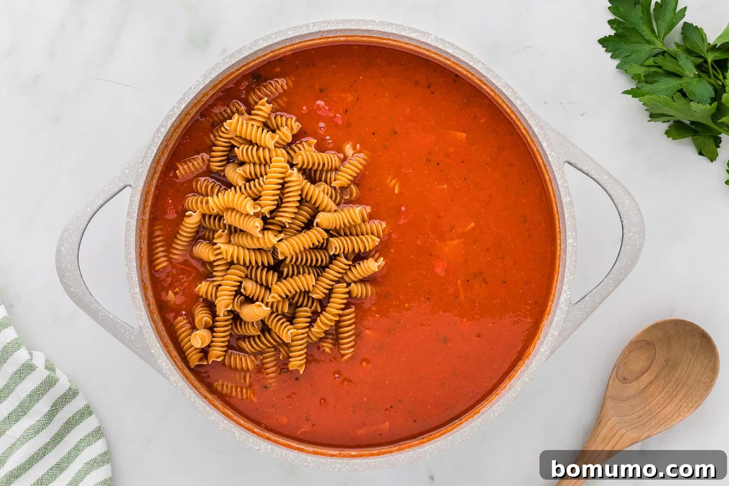 Uncooked rotini pasta being added on top of a simmering tomato sauce and pepperoni mixture in a large skillet.