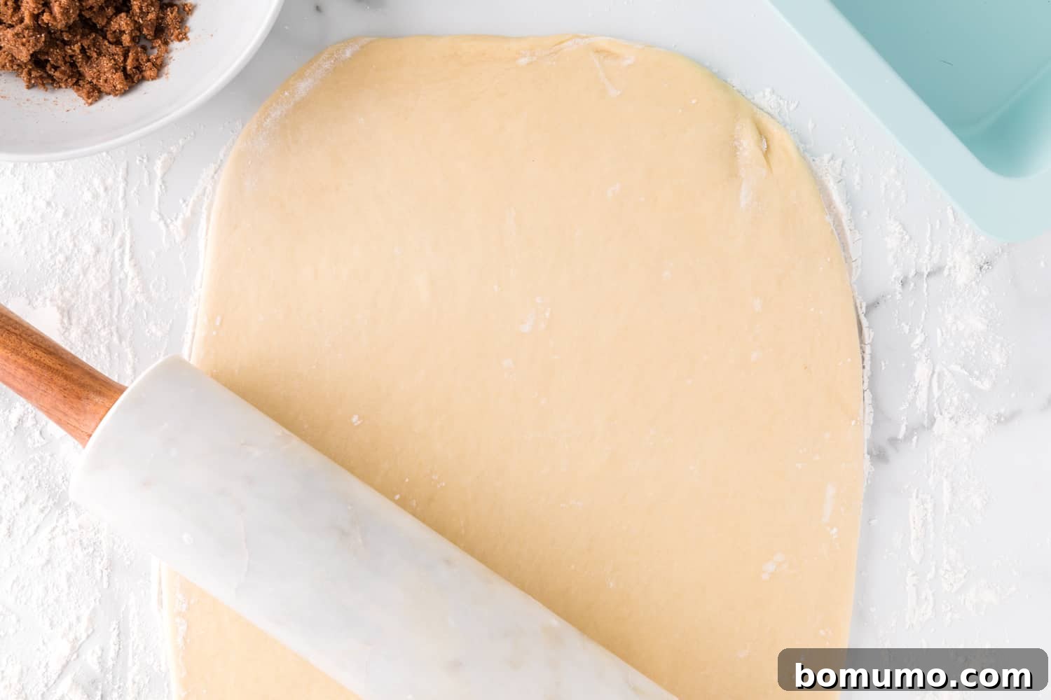 Bread dough being rolled out into a large rectangle on a floured surface.