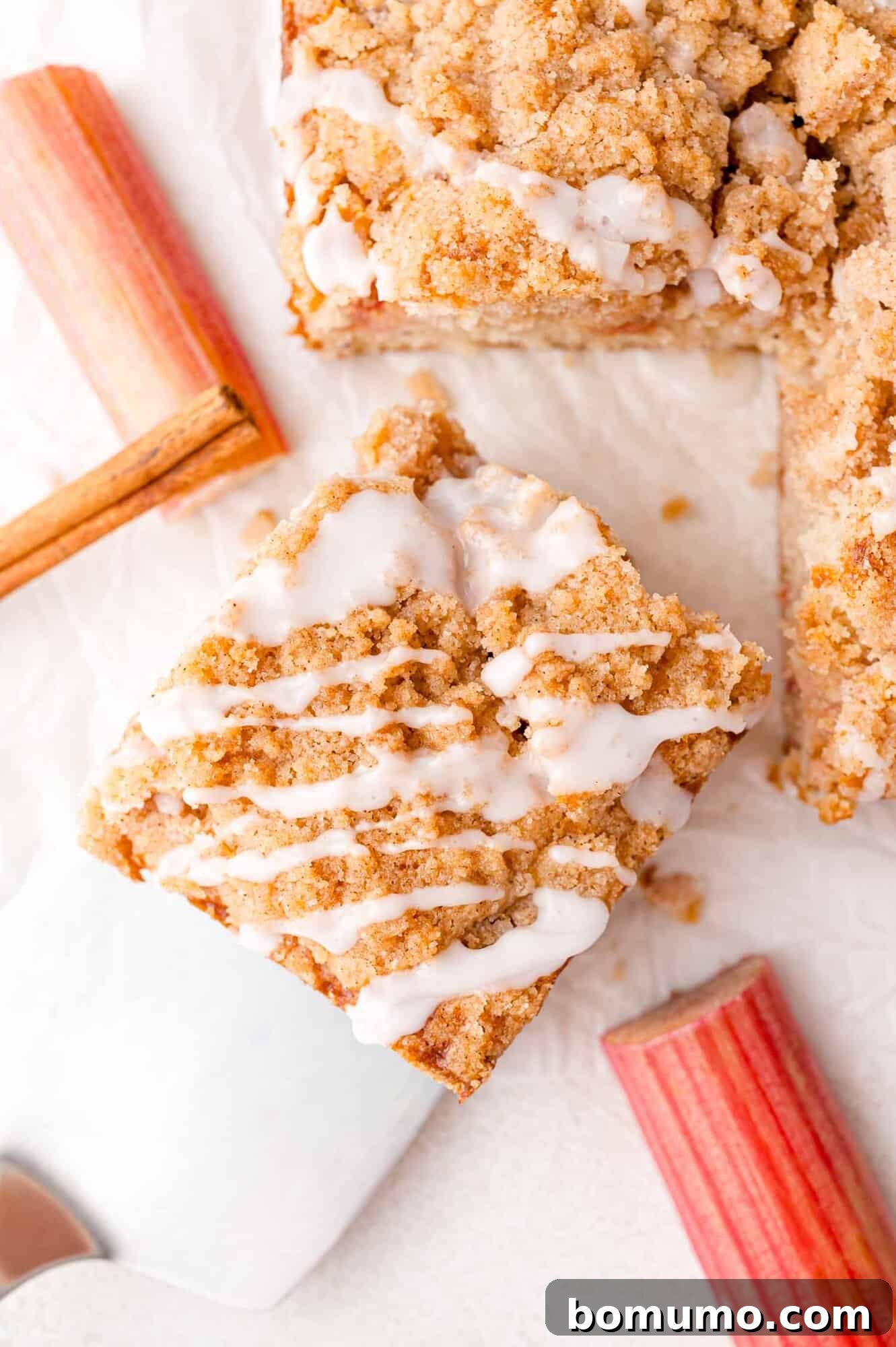 Tangy Rhubarb Cake 2 A square slice of crumb-topped coffee cake with a delicate icing drizzle, artfully arranged on a white surface with fresh rhubarb stalks beside it.