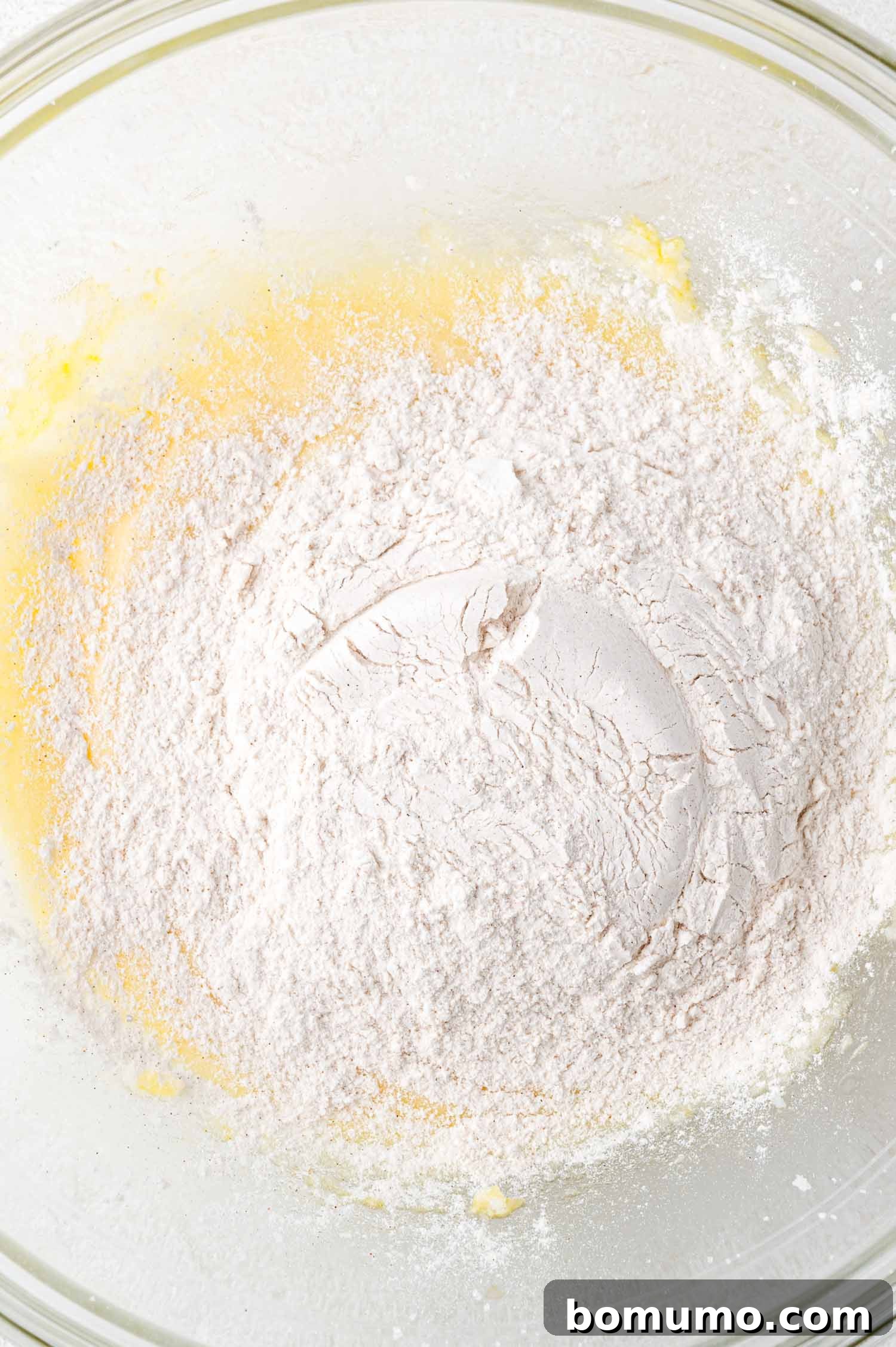 Tangy Rhubarb Cake 12 A glass mixing bowl holds a bright yellow, partially mixed cake batter with a layer of white all-purpose flour scattered on top, ready for the next mixing stage.