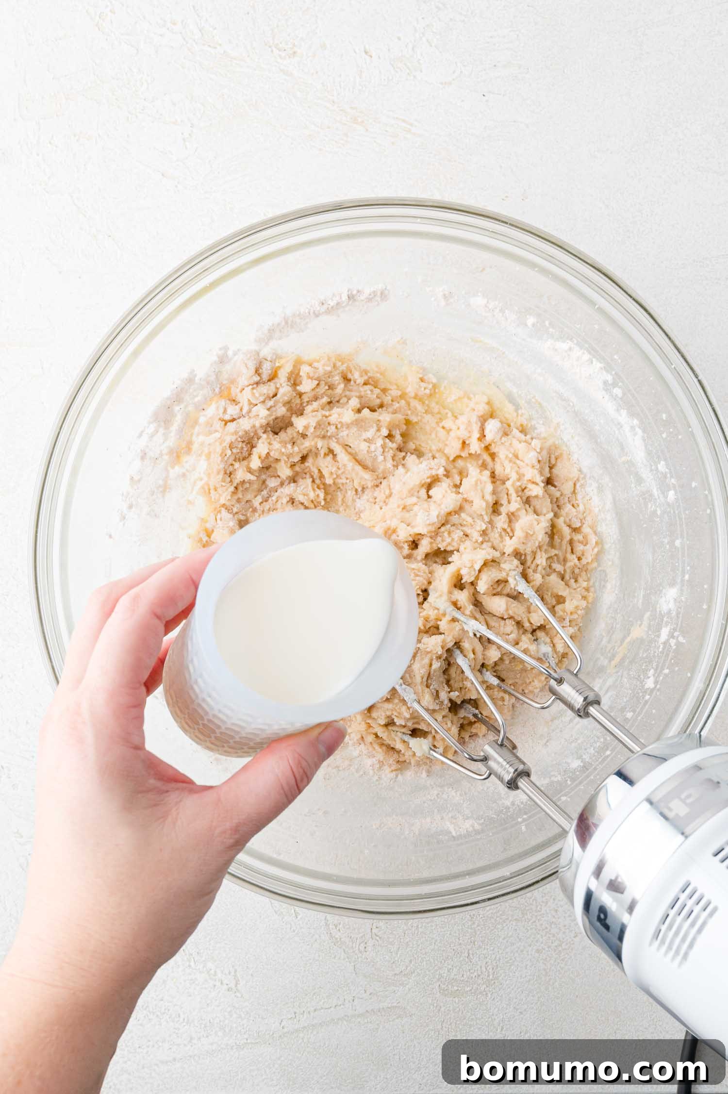 Tangy Rhubarb Cake 13 A hand holds a measuring cup, pouring fresh milk into a mixing bowl containing a creamy cake batter, with an electric mixer resting nearby.