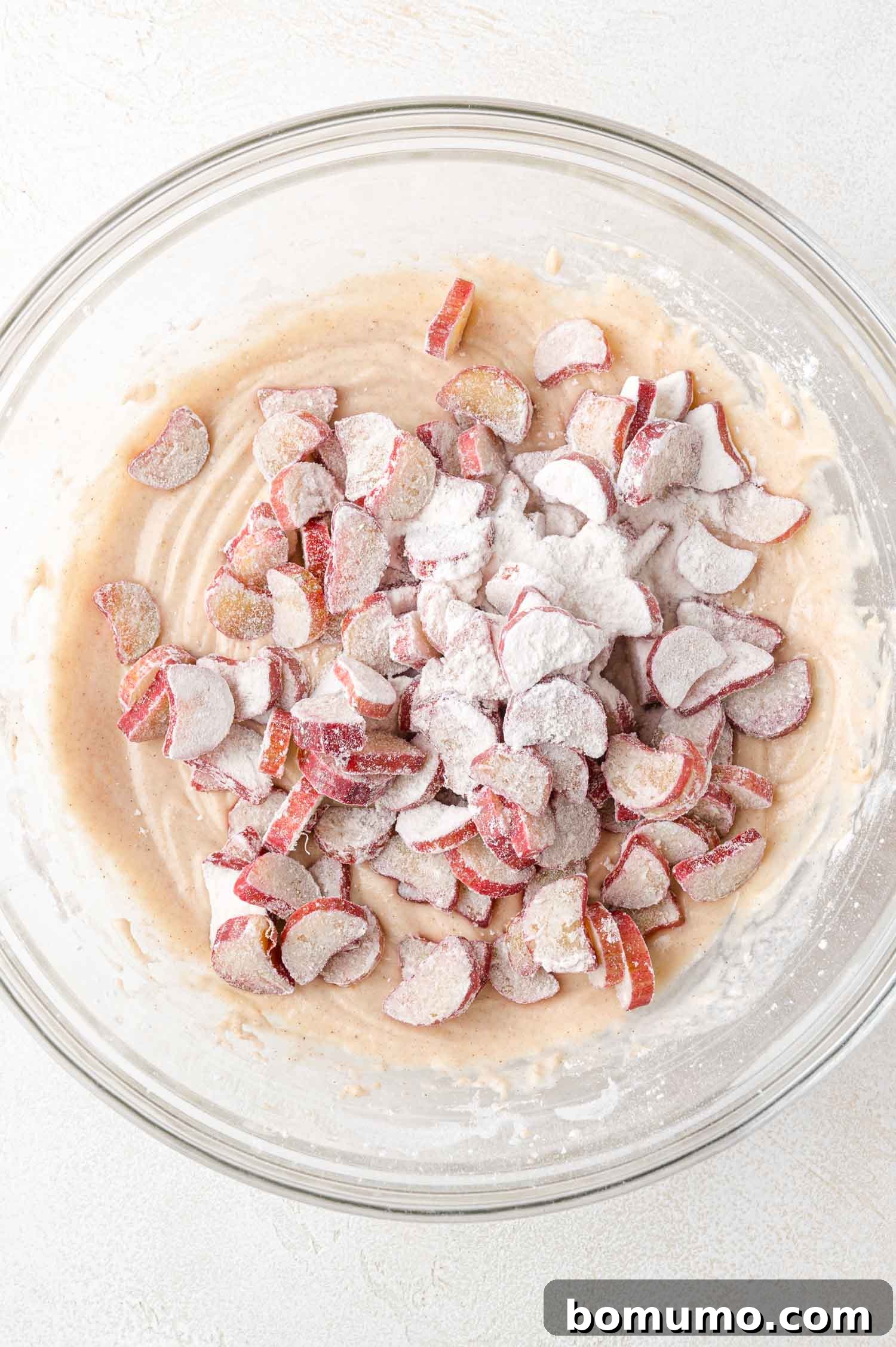 Tangy Rhubarb Cake 15 A glass bowl containing a rich, creamy cake batter meticulously mixed with vibrant, sliced rhubarb pieces, lightly coated in flour, resting on a light-colored surface.