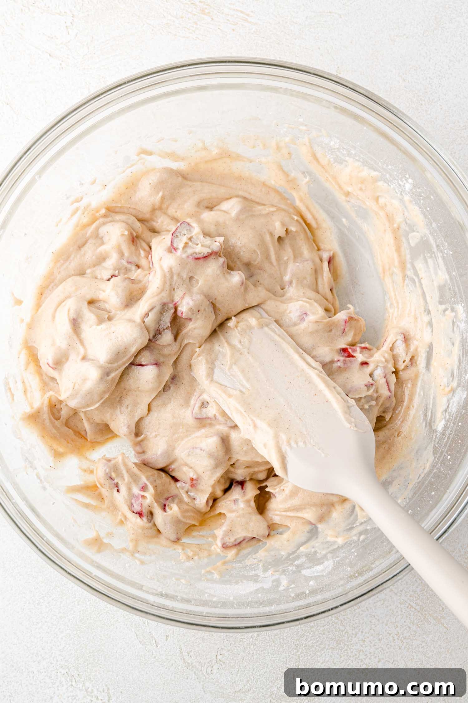 Tangy Rhubarb Cake 16 A clear glass bowl filled with thick, beige cake batter, generously mixed with bright red rhubarb pieces, being gently folded with a white spatula on a light surface.