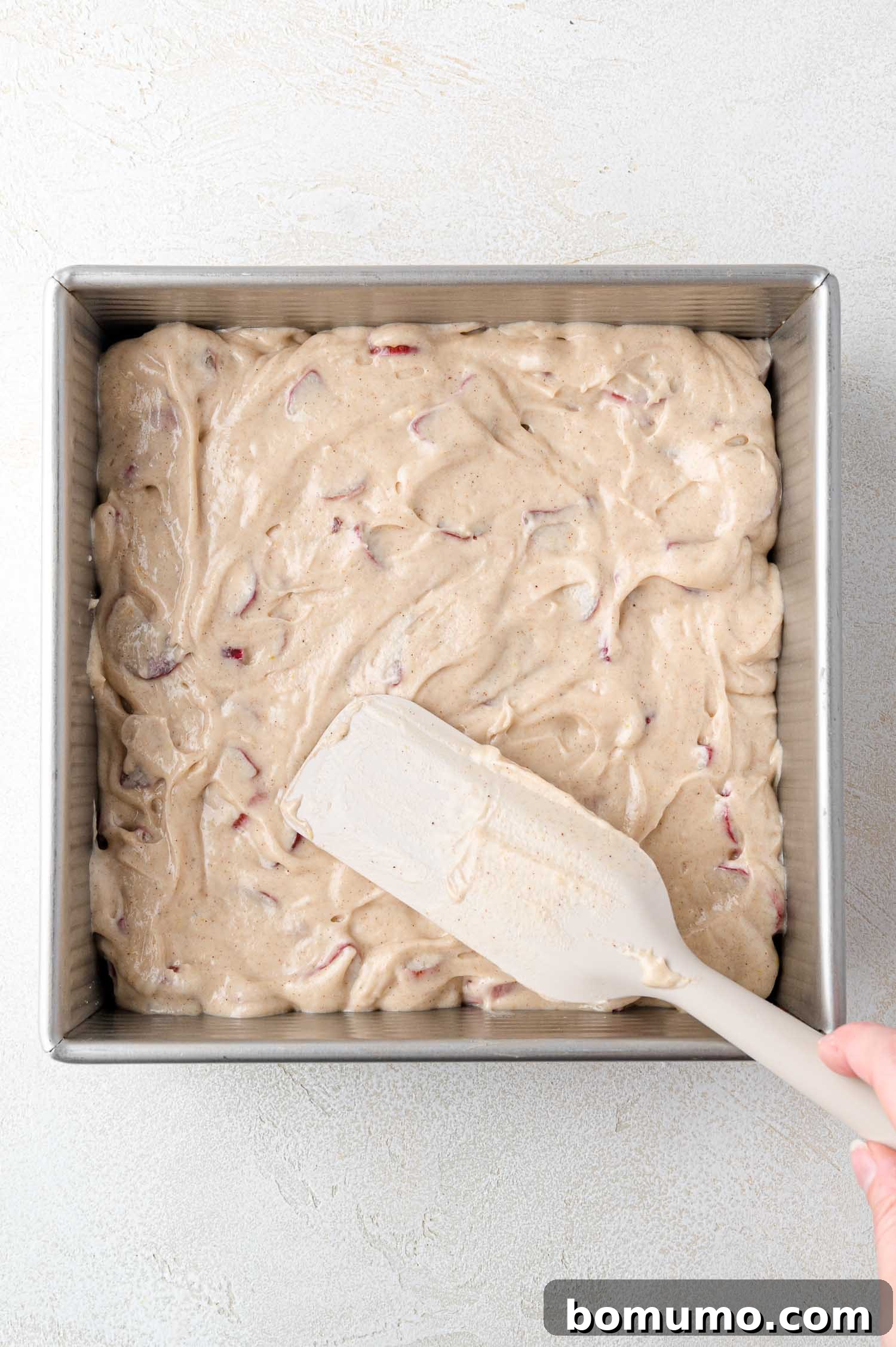 Tangy Rhubarb Cake 17 A hand skillfully uses a spatula to spread creamy cake batter, visibly mixed with vibrant rhubarb pieces, evenly across the bottom of a square metal baking pan.