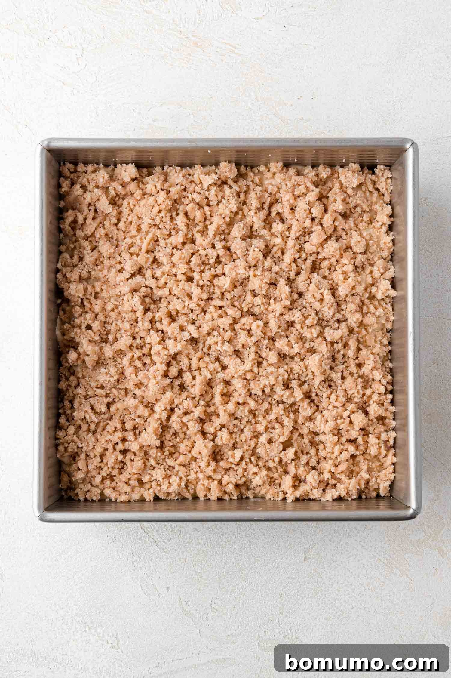Tangy Rhubarb Cake 18 A square metal baking pan holds an unbaked cake, topped with a generous, even layer of golden-brown crumb topping, resting on a light-colored surface, ready for the oven.