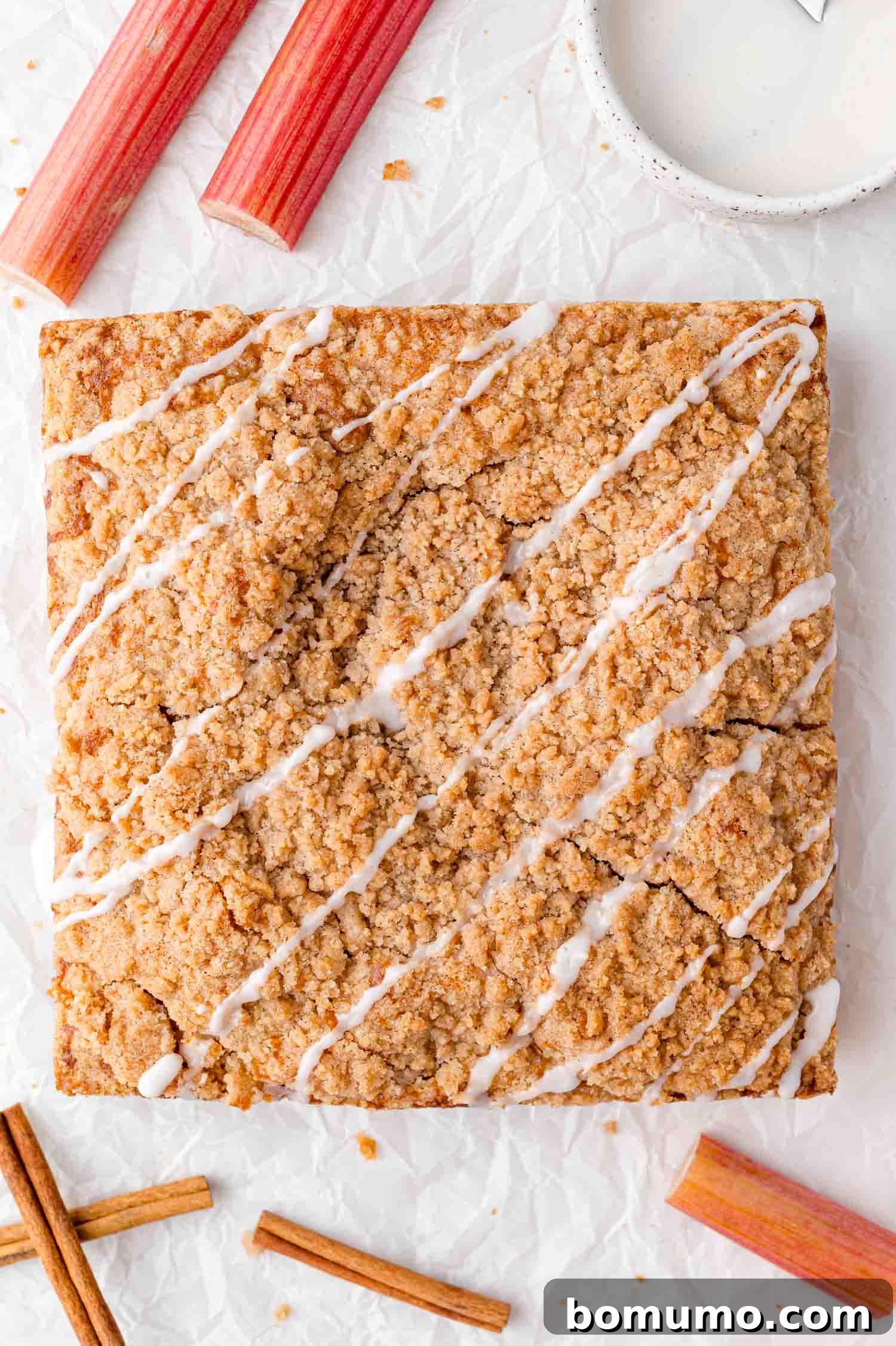 Tangy Rhubarb Cake 19 A beautifully baked square crumb cake, adorned with a delicate white icing drizzle, elegantly placed on crumpled parchment paper, surrounded by fresh rhubarb stalks, aromatic cinnamon sticks, and a small bowl of glaze.