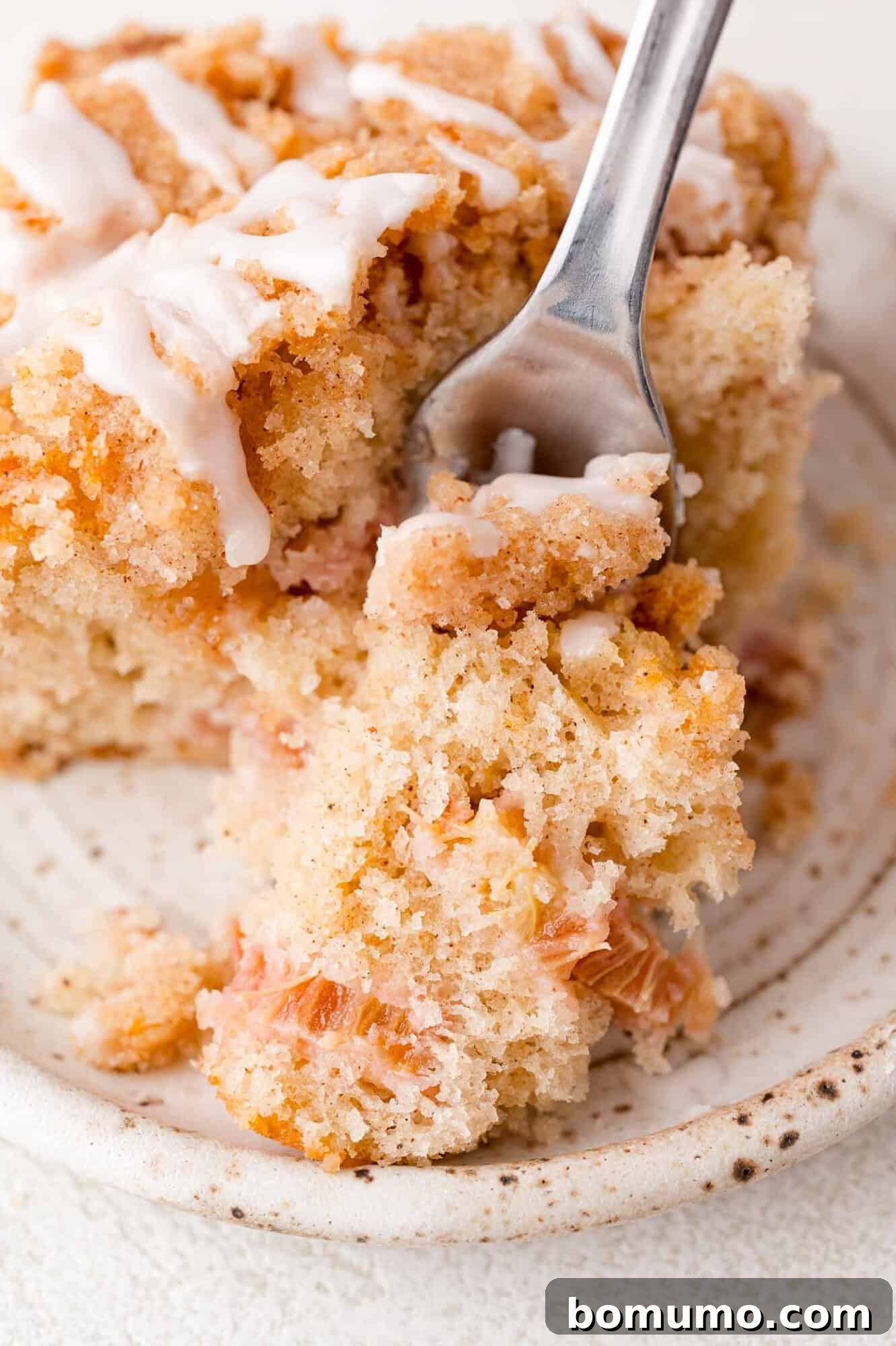 Tangy Rhubarb Cake 3 A close-up shot of a fork gently cutting into a slice of crumbly, iced coffee cake, resting on an elegant white ceramic plate.