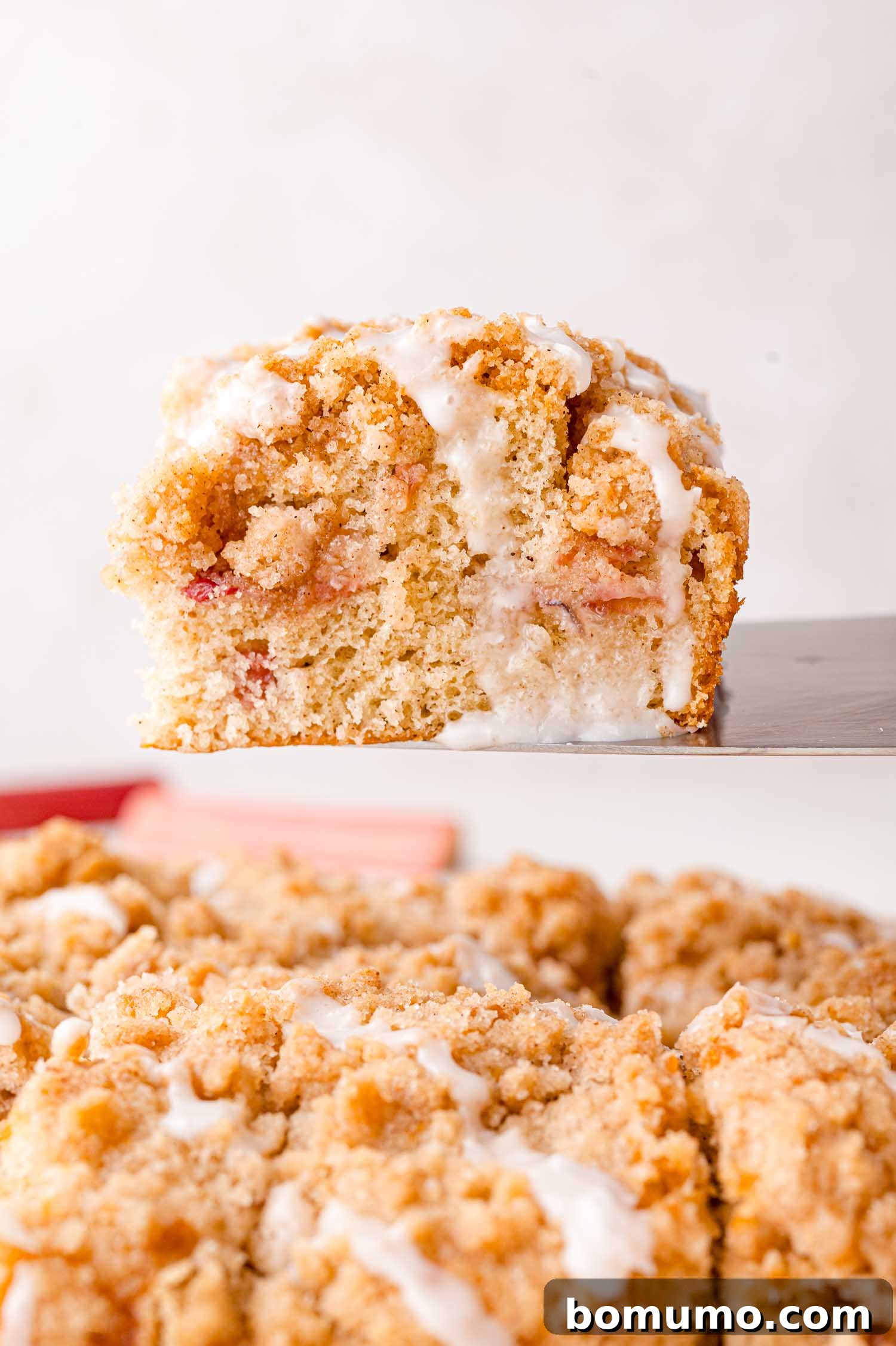 Tangy Rhubarb Cake 20 A close-up view of a perfectly sliced piece of crumb cake, featuring a generous drizzle of white icing, meticulously lifted above a pan filled with more delectable cake pieces.